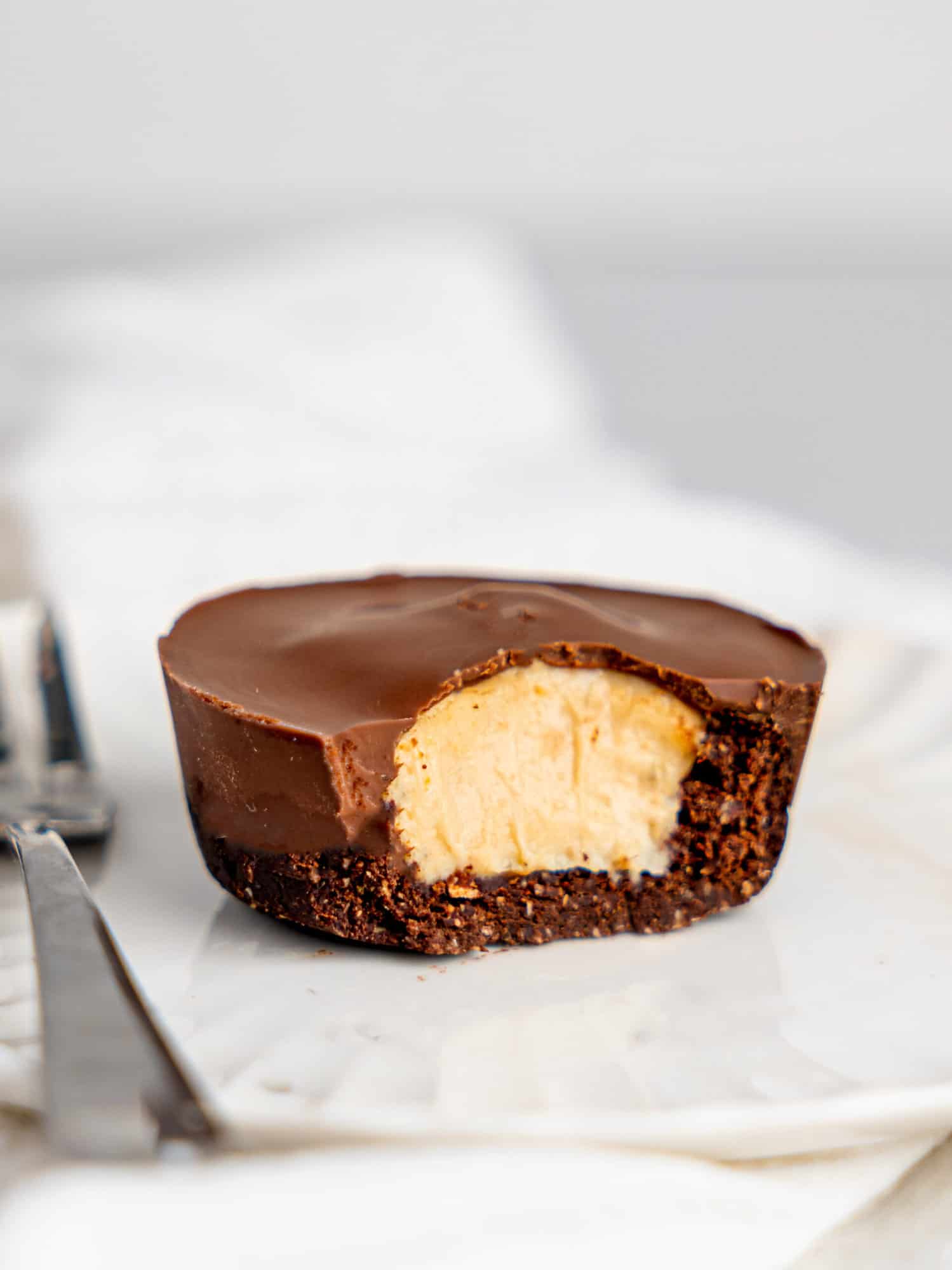 A peanut butter cup cheesecake on a small white plate, a cake fork to the left. A bite i missing from the cheesecake reavealing the creamy filling.