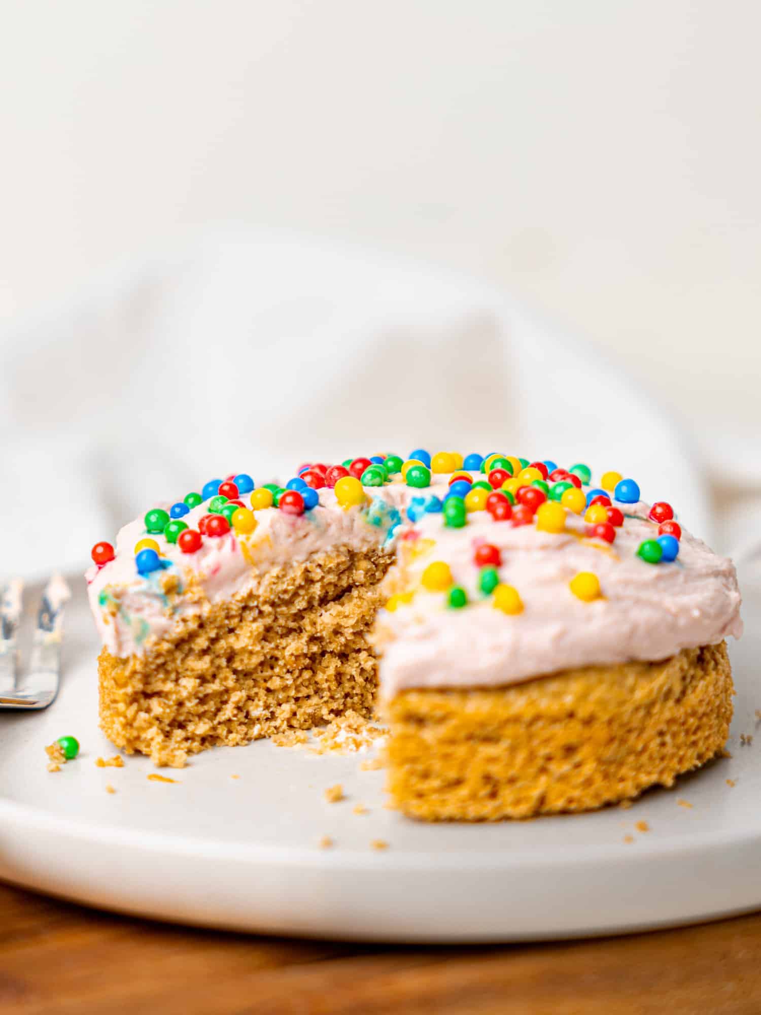 A single-serve birthday with pink protein frosting and sprinkles on a plate. A section is missing from the cake and there is a cake fork on the plate.