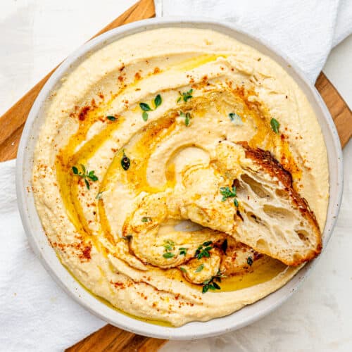 Creamy Cottage Cheese Hummus with a drizzle of olive oil spread out on a plate. There is a piece of foccaccia dipped into the hummus.