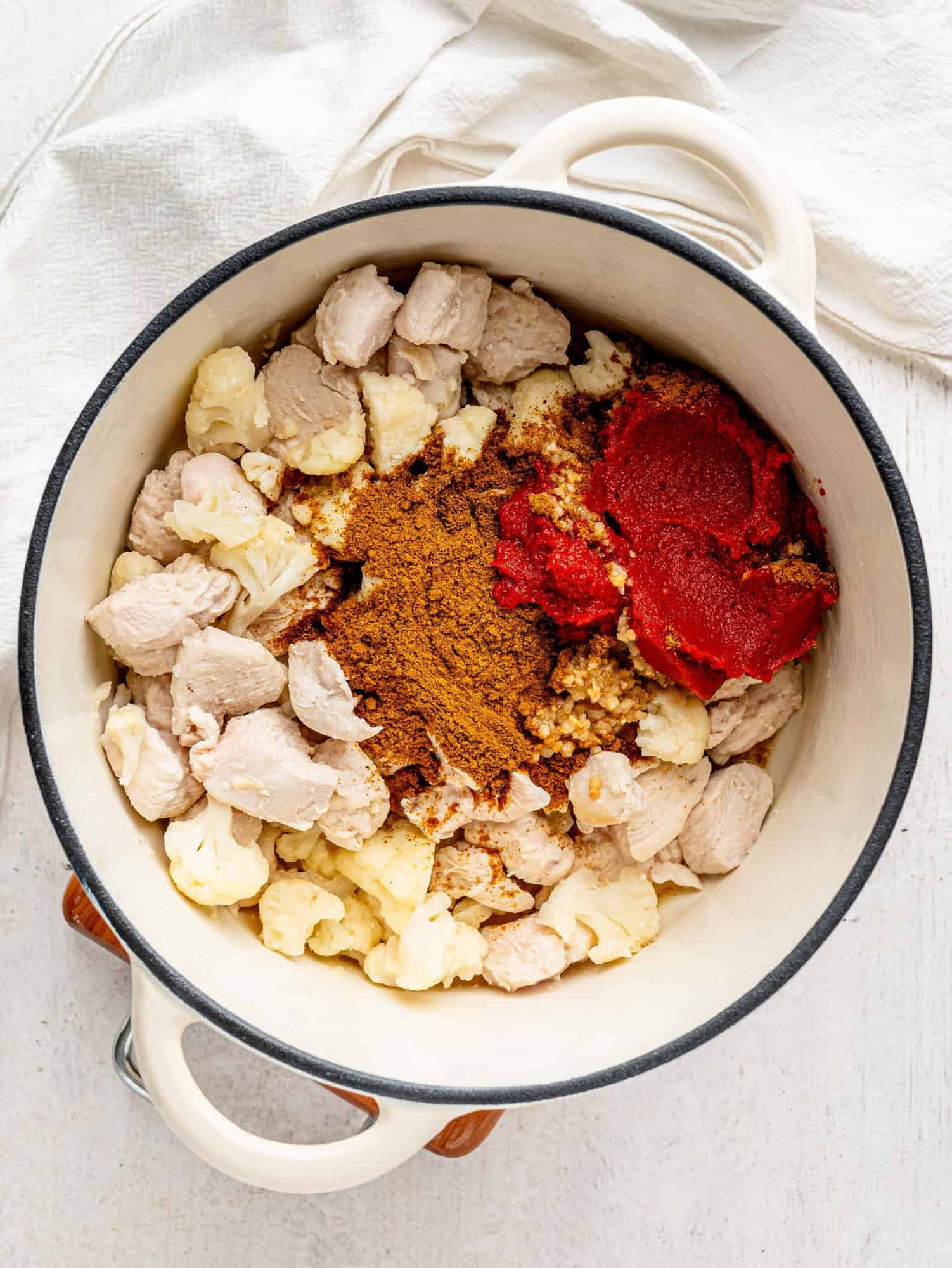 Cooked chicken and cauliflower, spices, tomato puree and garlic and ginger in a white pot.