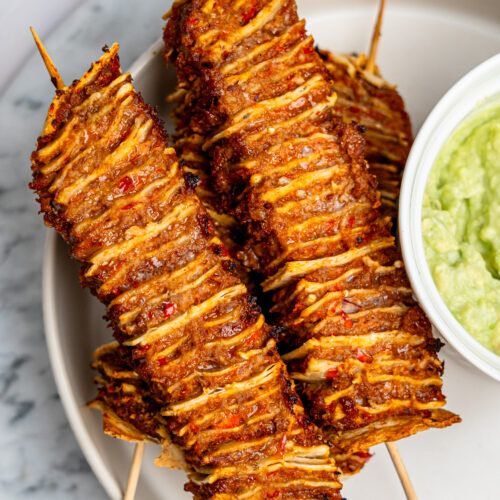 Four tortilla kebab skewers on a plate with a bowl of guacamole.