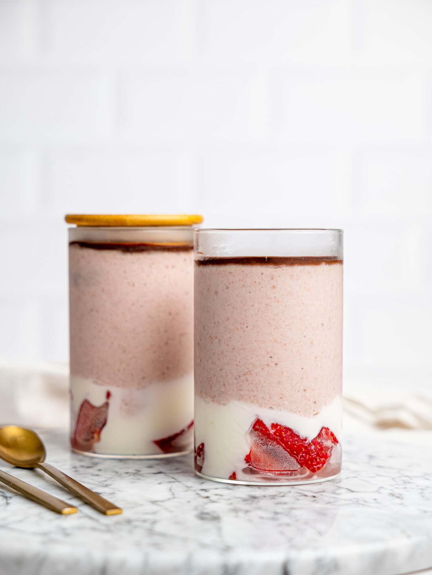 Two glasses of layered strawberry overnight oats on a marble surface, with strawberries and yogurt at the base, a creamy pink blended oat layer, and a thin chocolate topping, with spoons beside them.