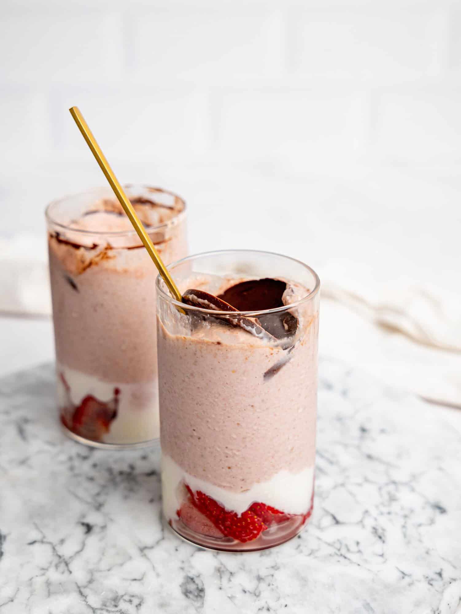 Two glasses of layered strawberry overnight oats on a marble surface, with strawberries and yogurt at the base, a creamy pink blended oat layer, and a chocolate topping, one with a spoon inserted.