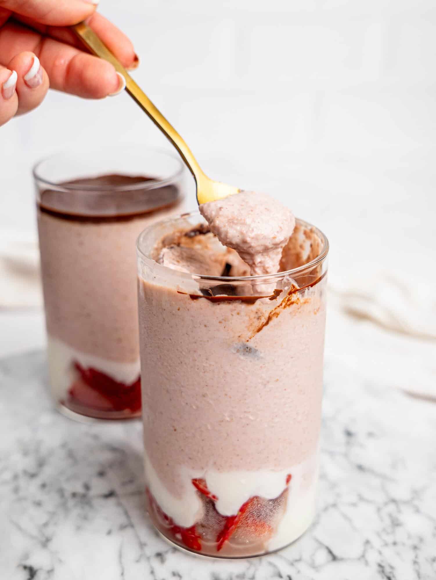 Close-up of a spoon lifting a creamy bite of strawberry blended overnight oats from a glass with visible chocolate layer and strawberry yogurt base.
