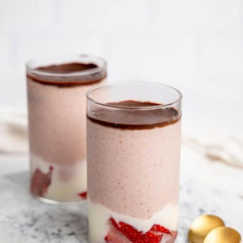 Two glasses of strawberry blended overnight oats on a marble surface with a smooth chocolate topping, with gold spoons placed beside them.