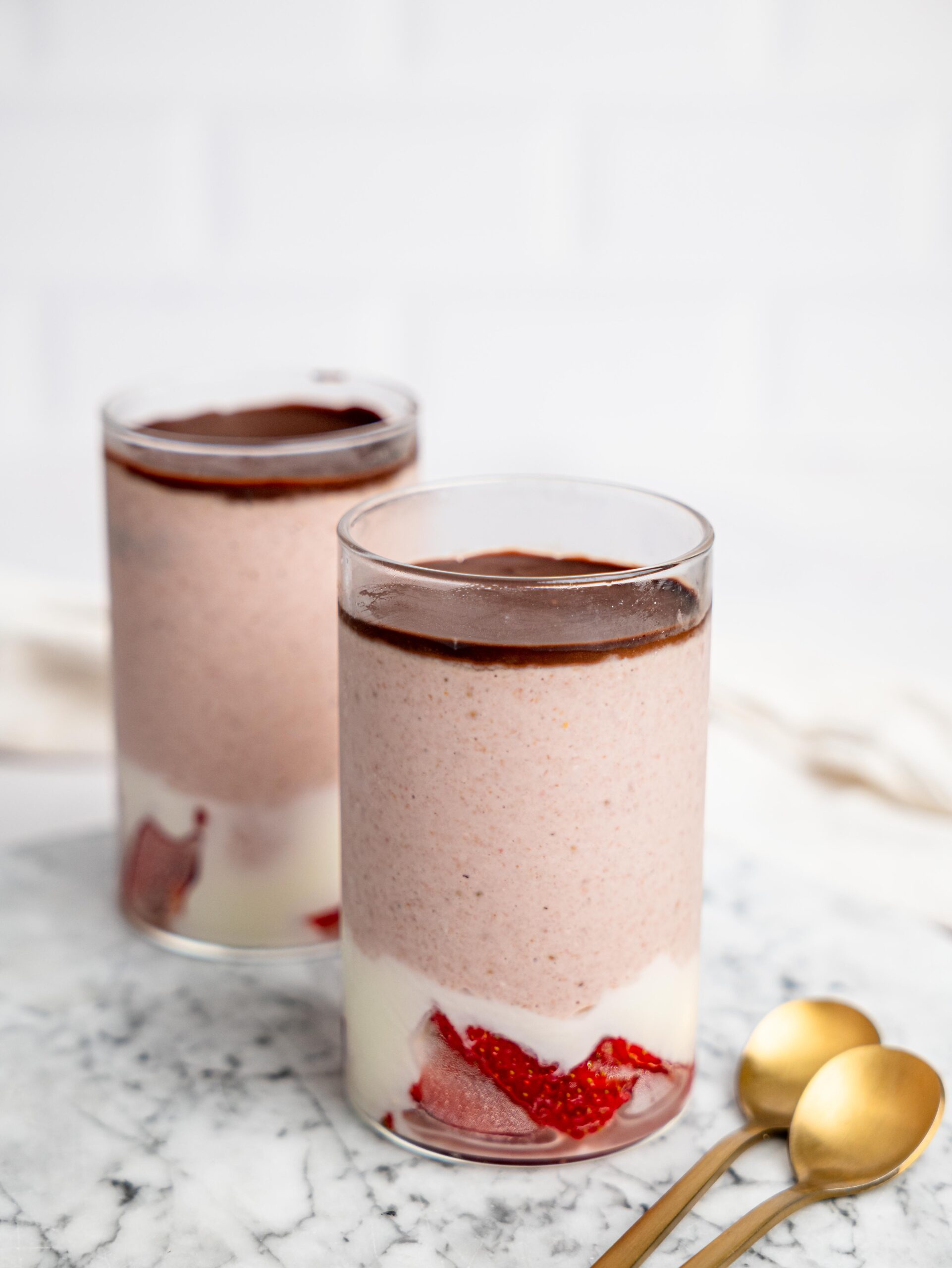 Two glasses of strawberry blended overnight oats on a marble surface with a smooth chocolate topping, with gold spoons placed beside them.