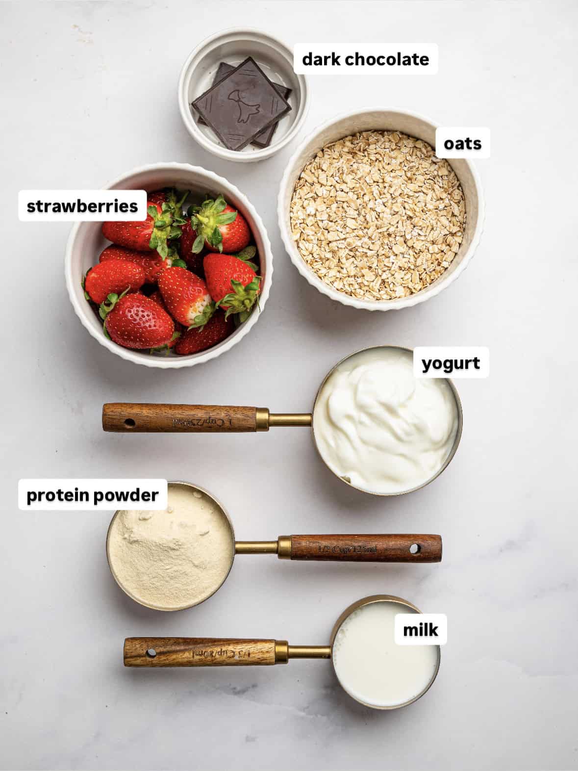 Flat lay of ingredients for blended overnight oats on a light surface, including fresh strawberries in a bowl, rolled oats, dark chocolate squares, yogurt, protein powder, and milk measured in wooden-handled cups.