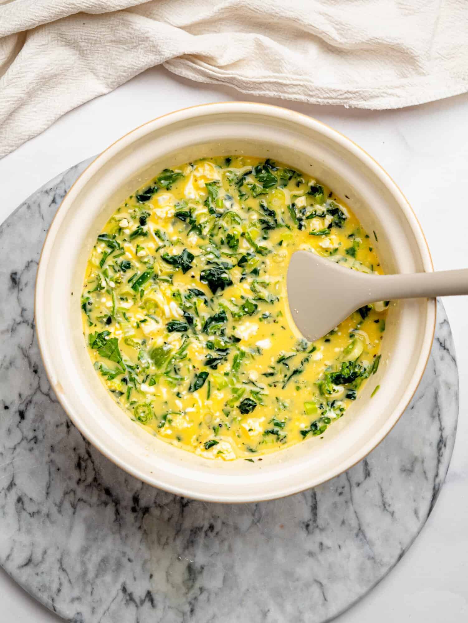 Egg mixture with chopped spinach, herbs, and feta in a bowl, partially mixed before baking.