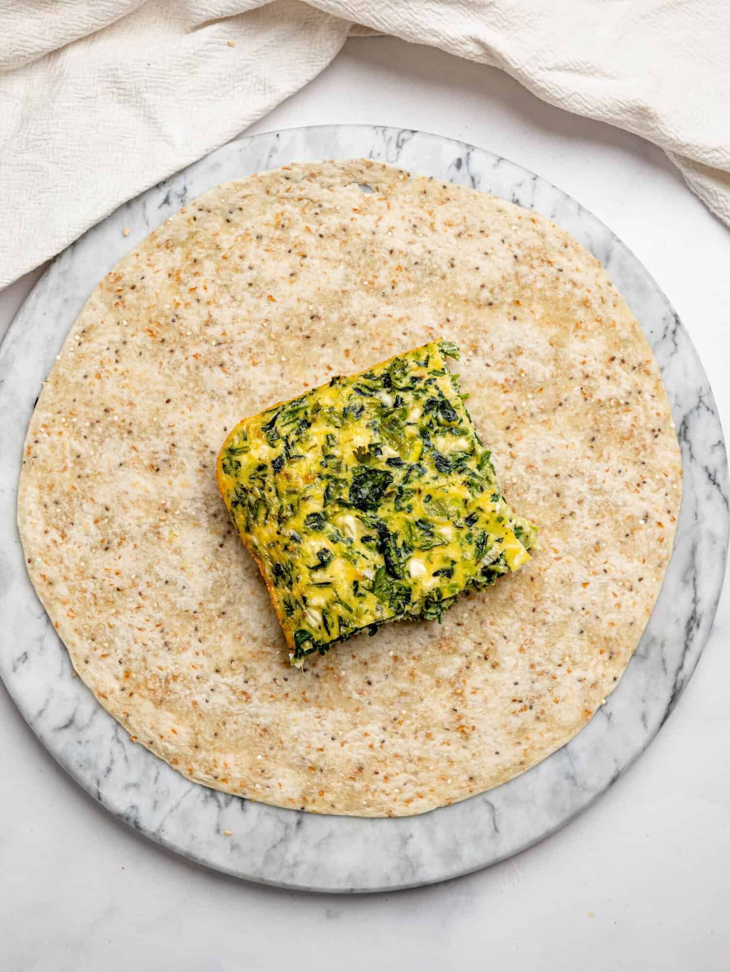 A square piece of baked spinach and feta egg filling placed in the center of a tortilla, ready to be wrapped.