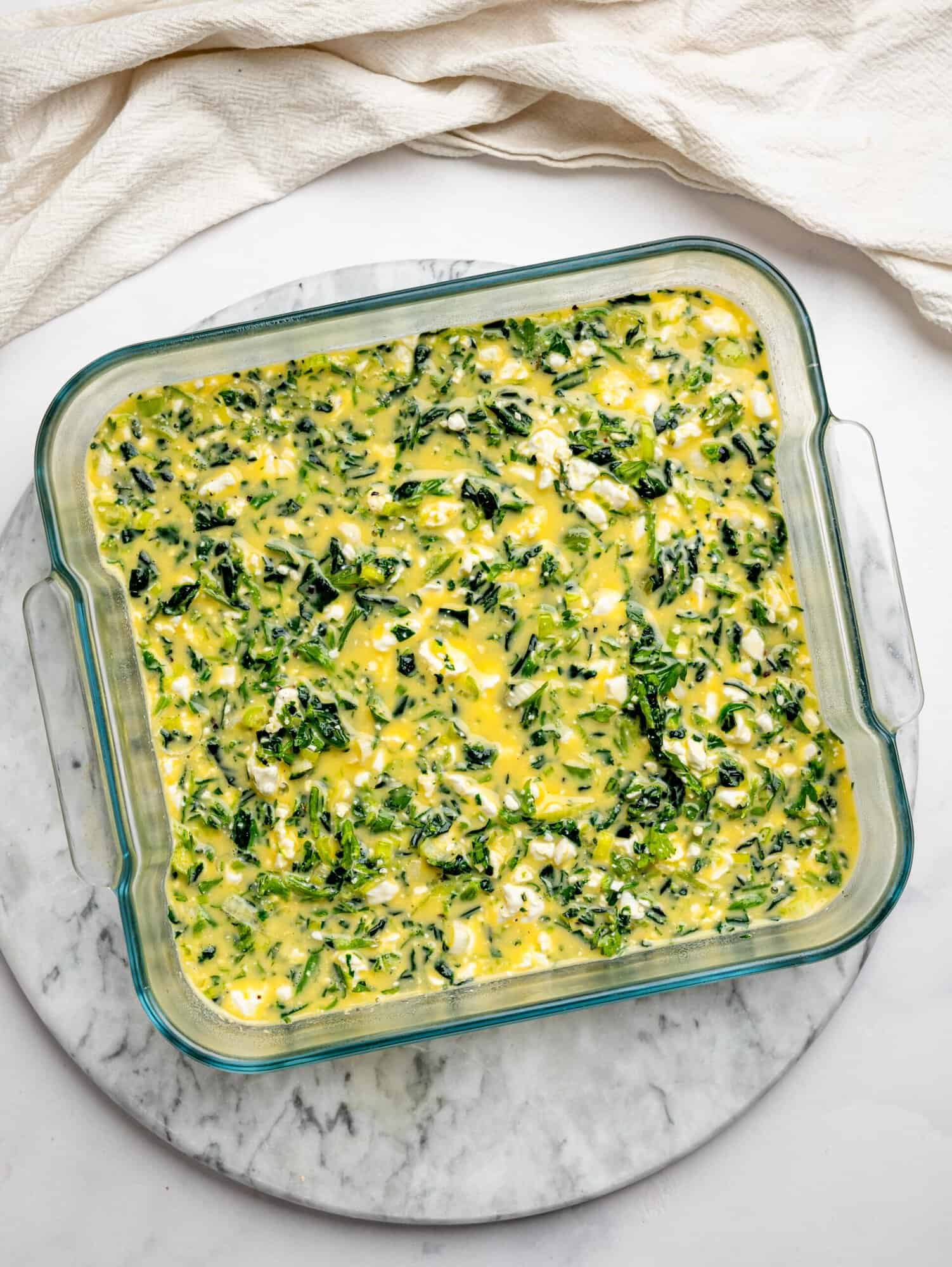 Unbaked egg mixture with spinach, herbs, and feta in a glass baking dish before cooking.