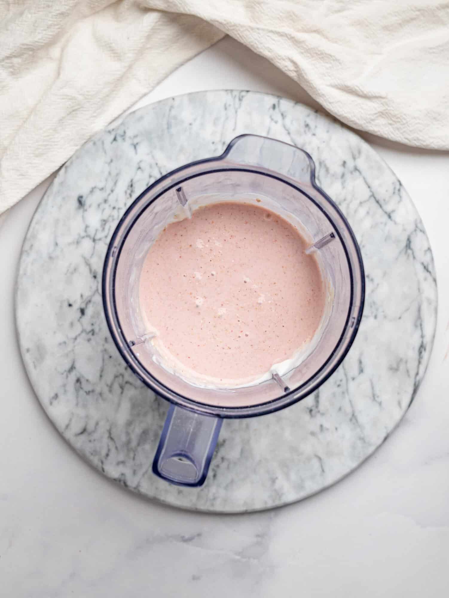 Top view of a blender jug on a marble board filled with smooth, pale pink blended mixture for strawberry overnight oats.