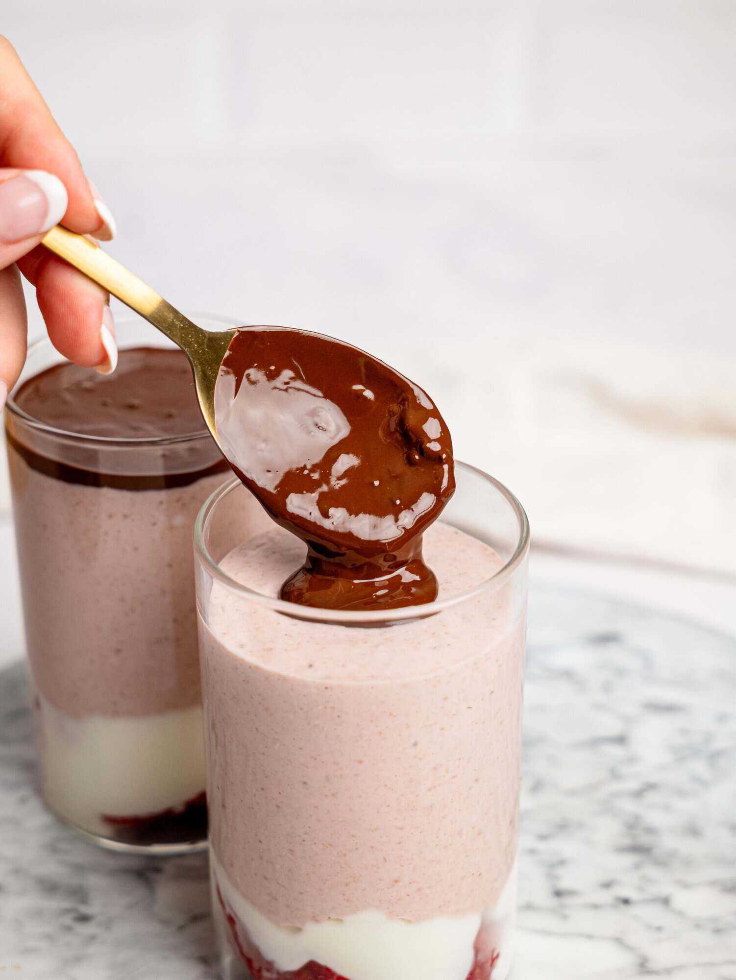 Close-up of a hand spooning melted chocolate over a glass of layered strawberry overnight oats with strawberries, and yogurt.
