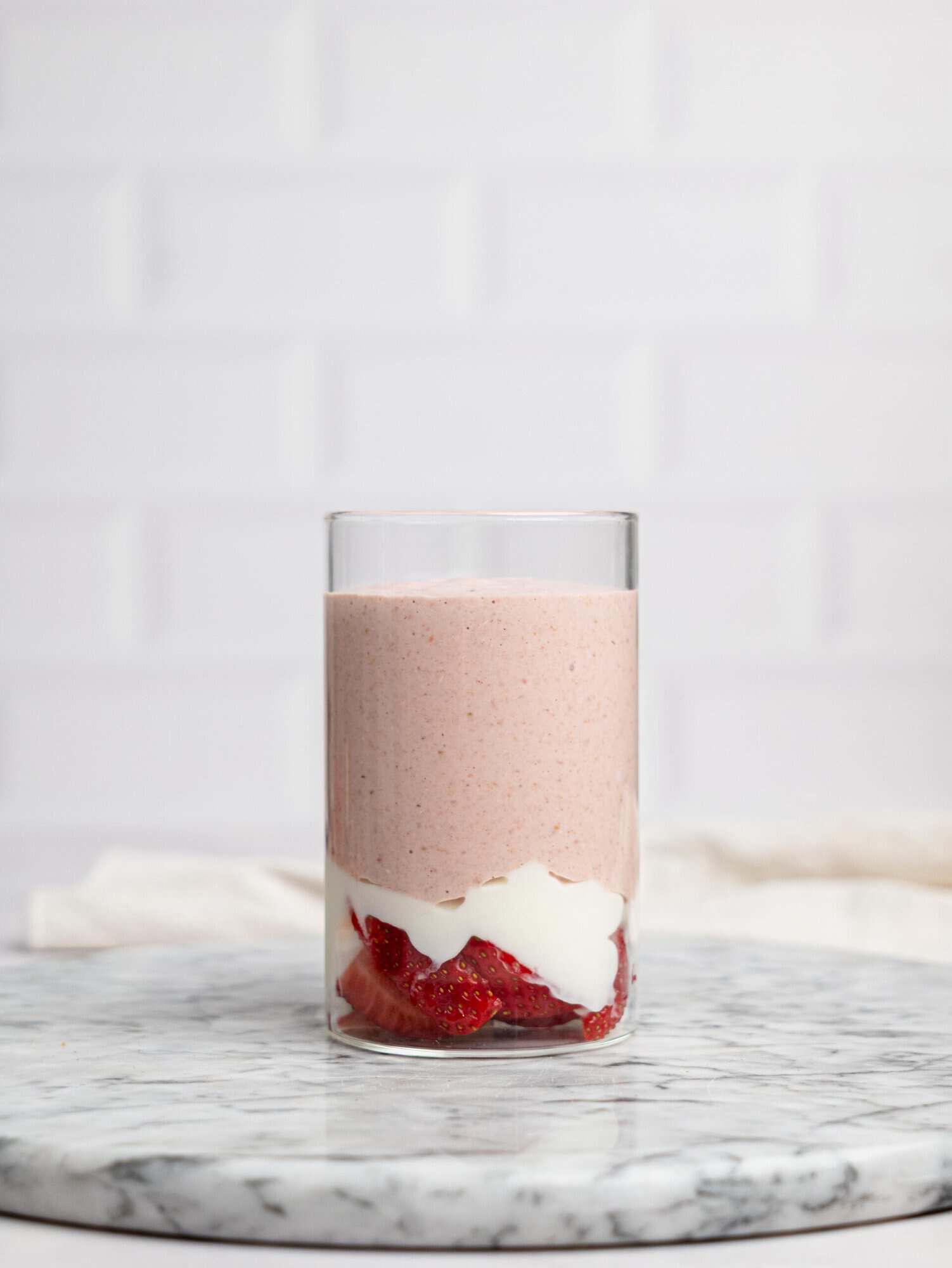 Side view of a clear glass on a marble surface layered with sliced strawberries, yogurt, and a smooth pink blended mixture for strawberry overnight oats.