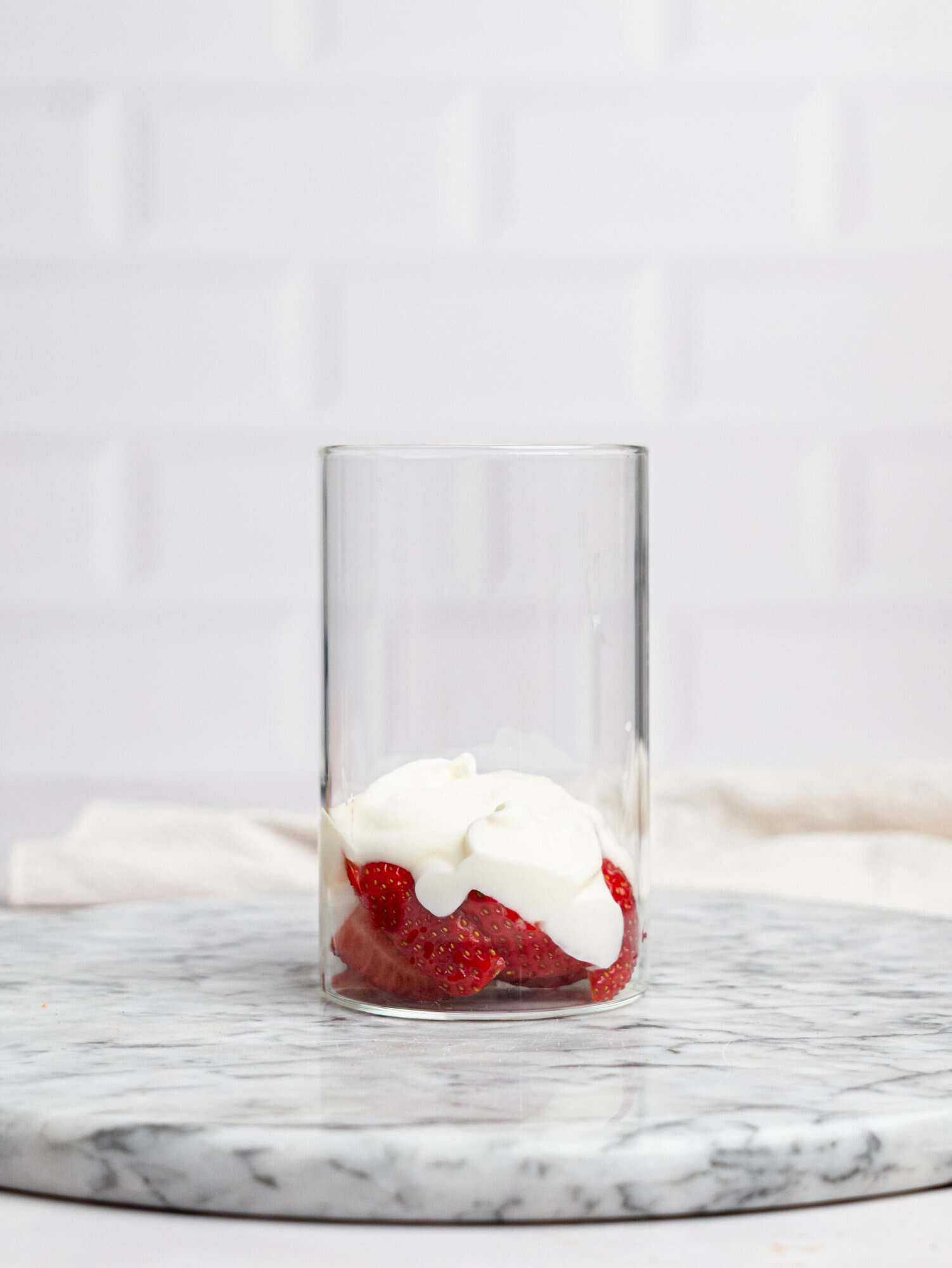 Side view of a clear glass on a marble surface with sliced strawberries and a dollop of yogurt at the bottom, ready for layered strawberry overnight oats.