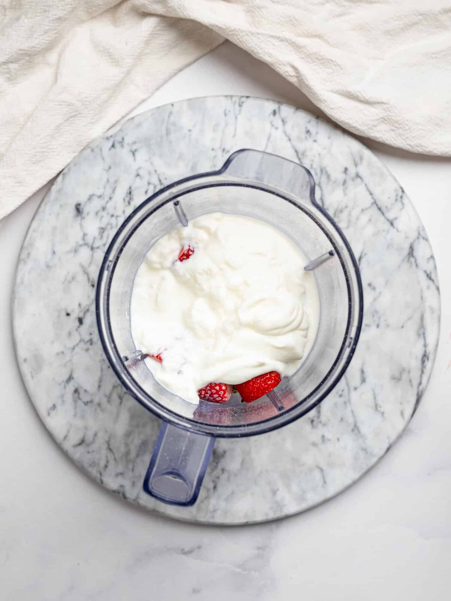 Top view of a blender jug on a marble board filled with ingredients for strawberry overnight oats, including yogurt and strawberries on top, with the remaining ingredients underneath ready to be blended.