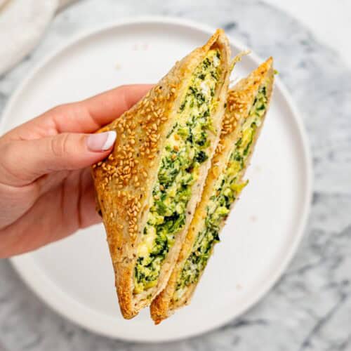 Hand holding a halved spanakopita egg wrap with a golden, sesame-crusted tortilla and a creamy spinach, egg, and feta filling visible inside.