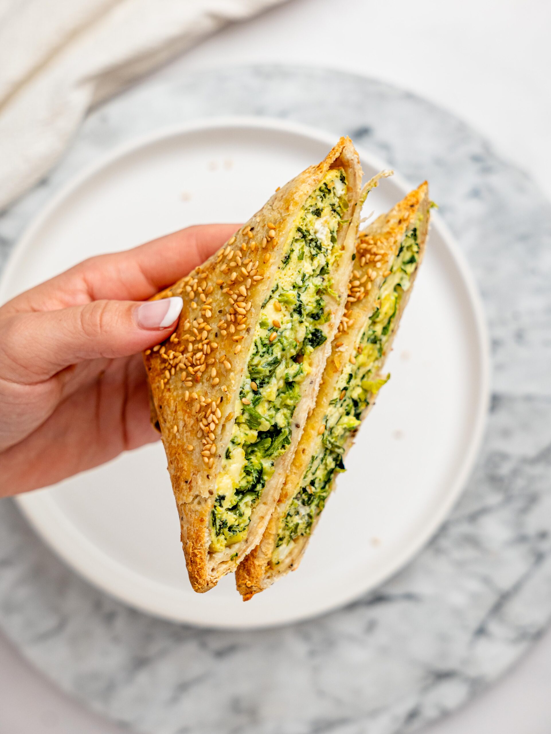 Hand holding a halved spanakopita egg wrap with a golden, sesame-crusted tortilla and a creamy spinach, egg, and feta filling visible inside.