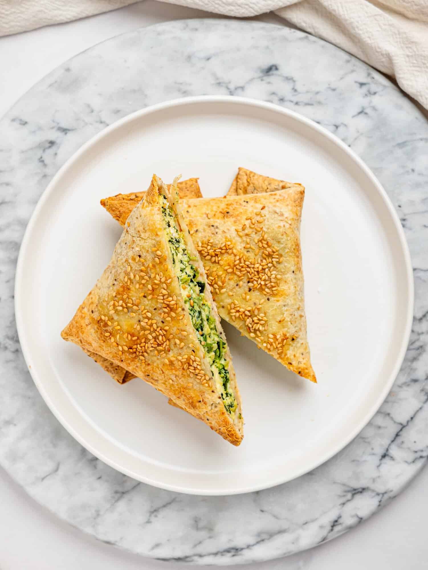 Overhead view of a spanakopita egg wrap on a white plate, lightly crisped and topped with sesame seeds.