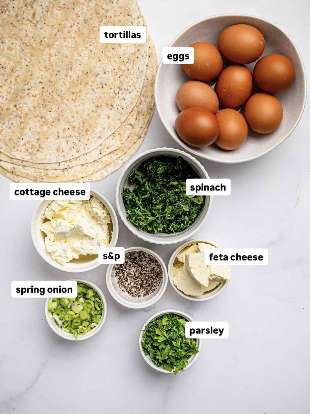 Ingredients laid out for spanakopita egg wraps, including tortillas, eggs, spinach, feta, herbs, and seasoning.