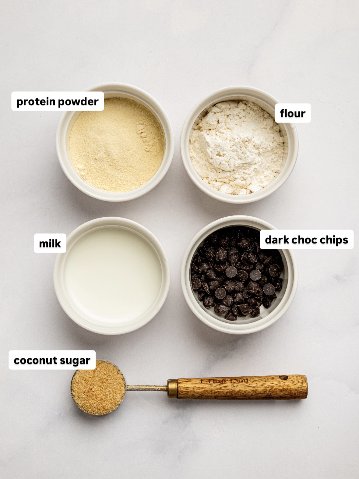 Small bowls of protein powder, flour, milk, dark chocolate chips, and coconut sugar arranged on a white surface for a healthy chocolate chip mug cake recipe.