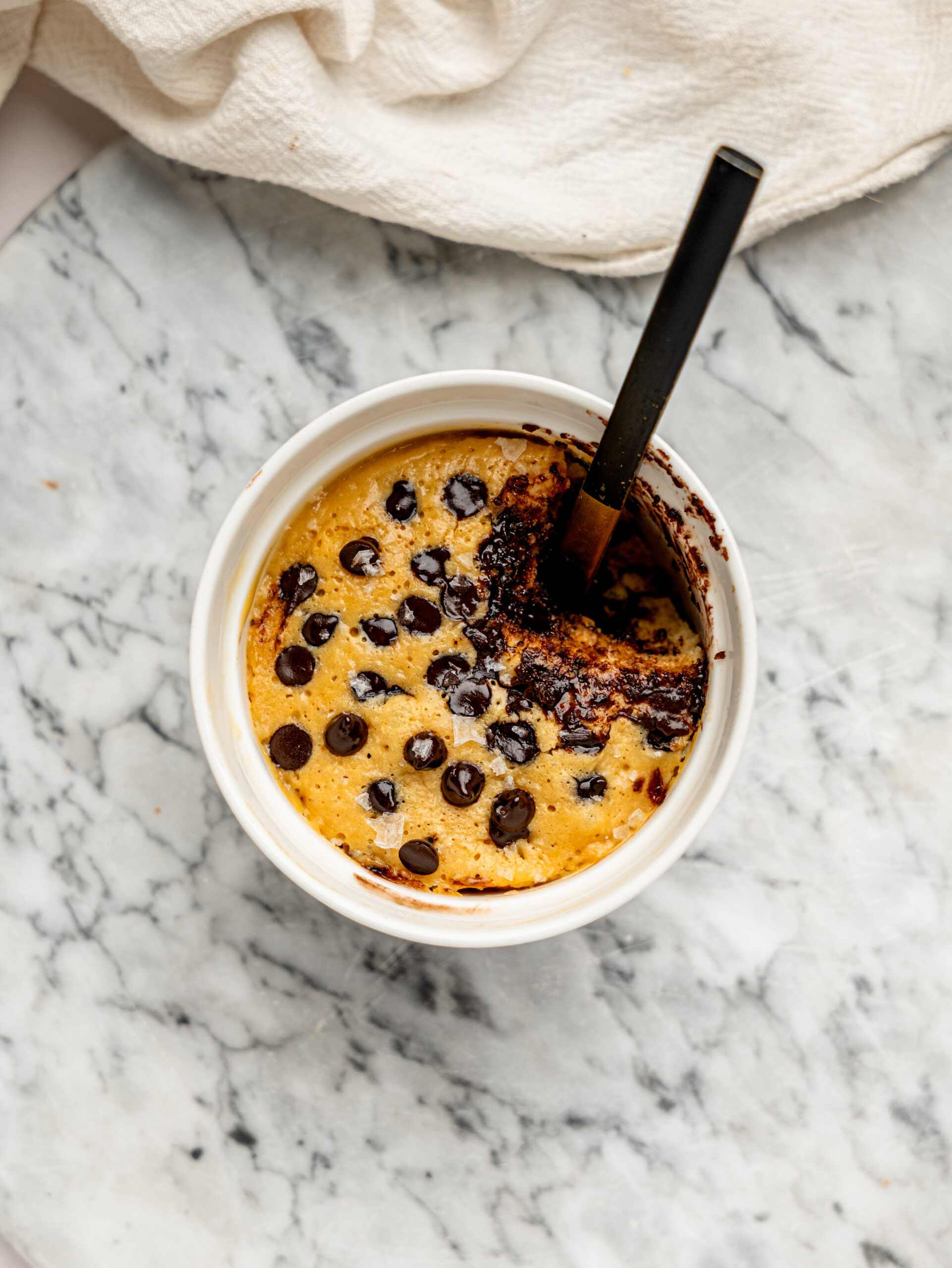 Freshly baked healthy chocolate chip mug cake in a white ramekin with melted chocolate chips and flaky sea salt on top.