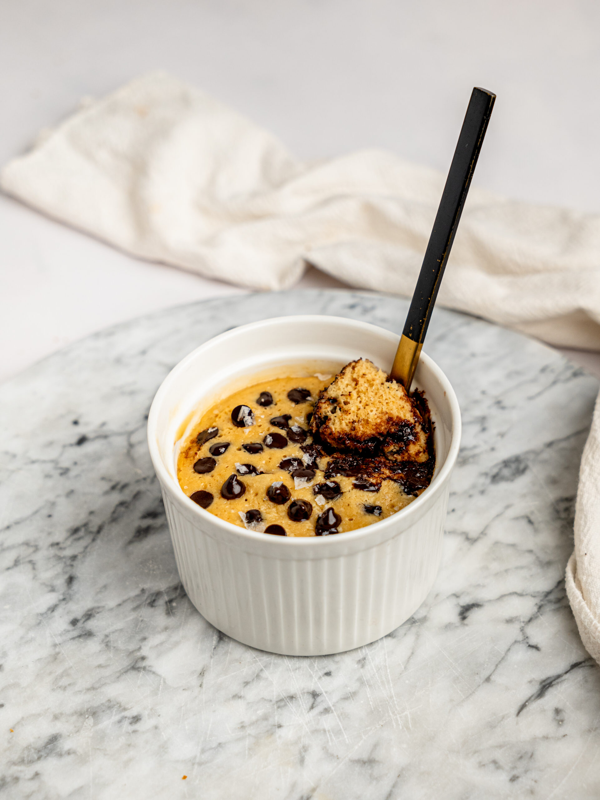 Healthy chocolate chip mug cake served in a white ramekin on a marble board with a spoon resting inside.