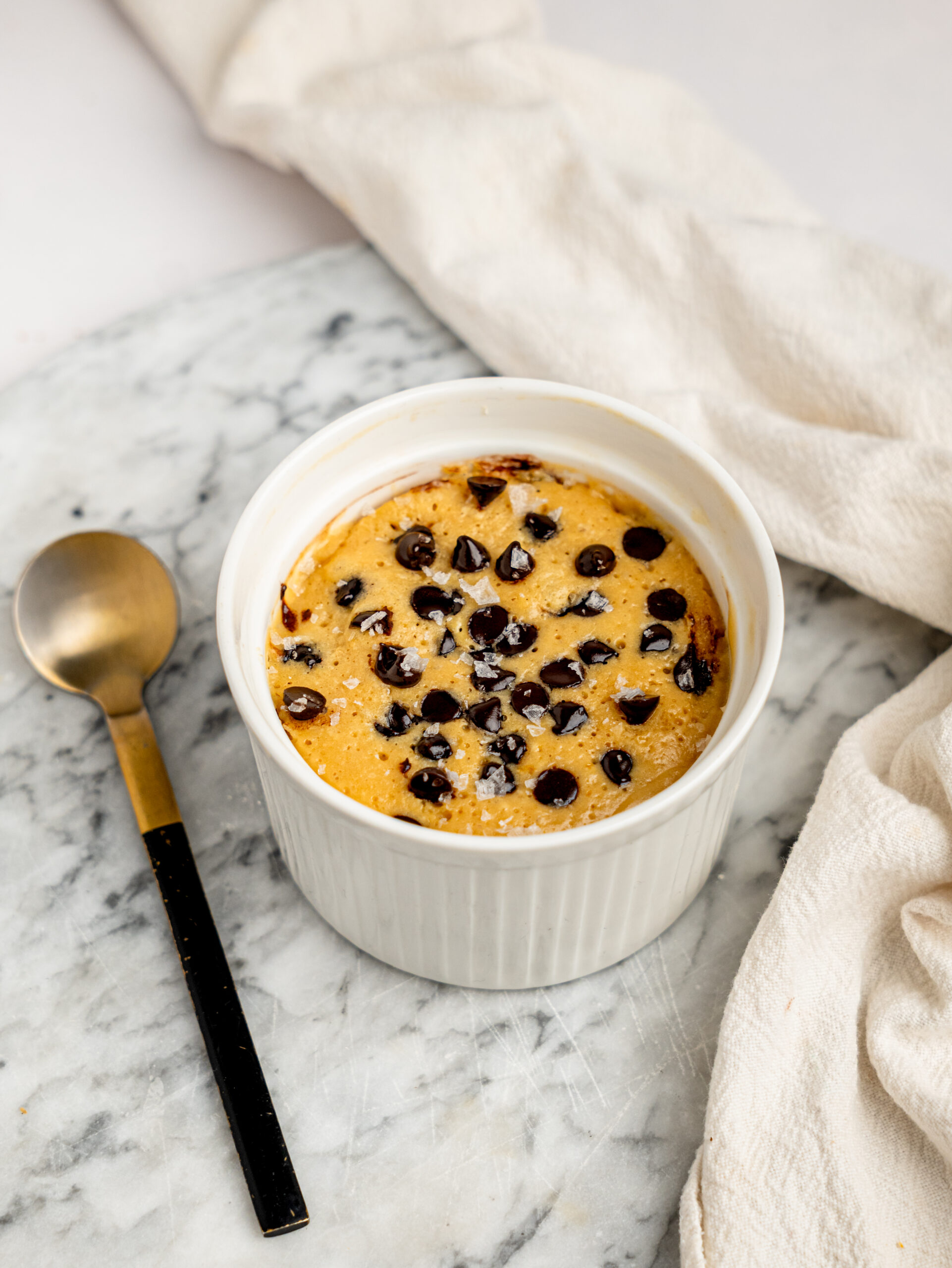 Overhead view of a healthy protein mug cake with melted chocolate chips in a white ramekin on a marble board.