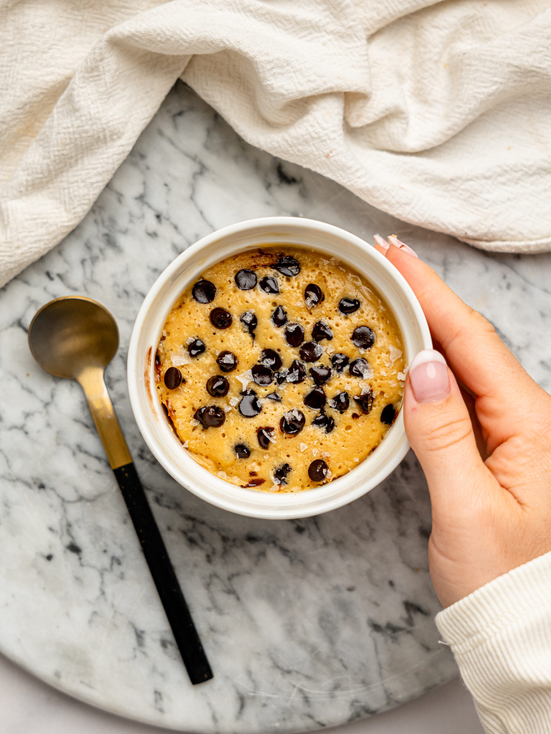 Hand holding a warm chocolate chip protein mug cake in a white ramekin beside a black and gold spoon.