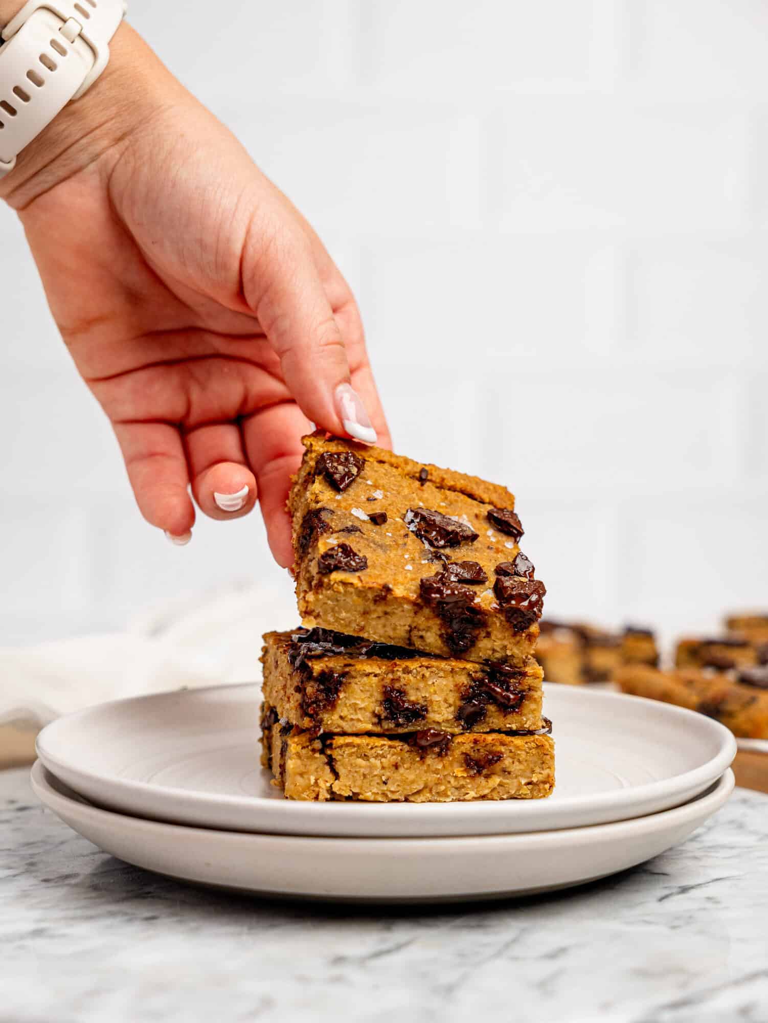 Three chickpea cookie bars stacked on top of each other on a white plate, a hand is picking up the one on top.