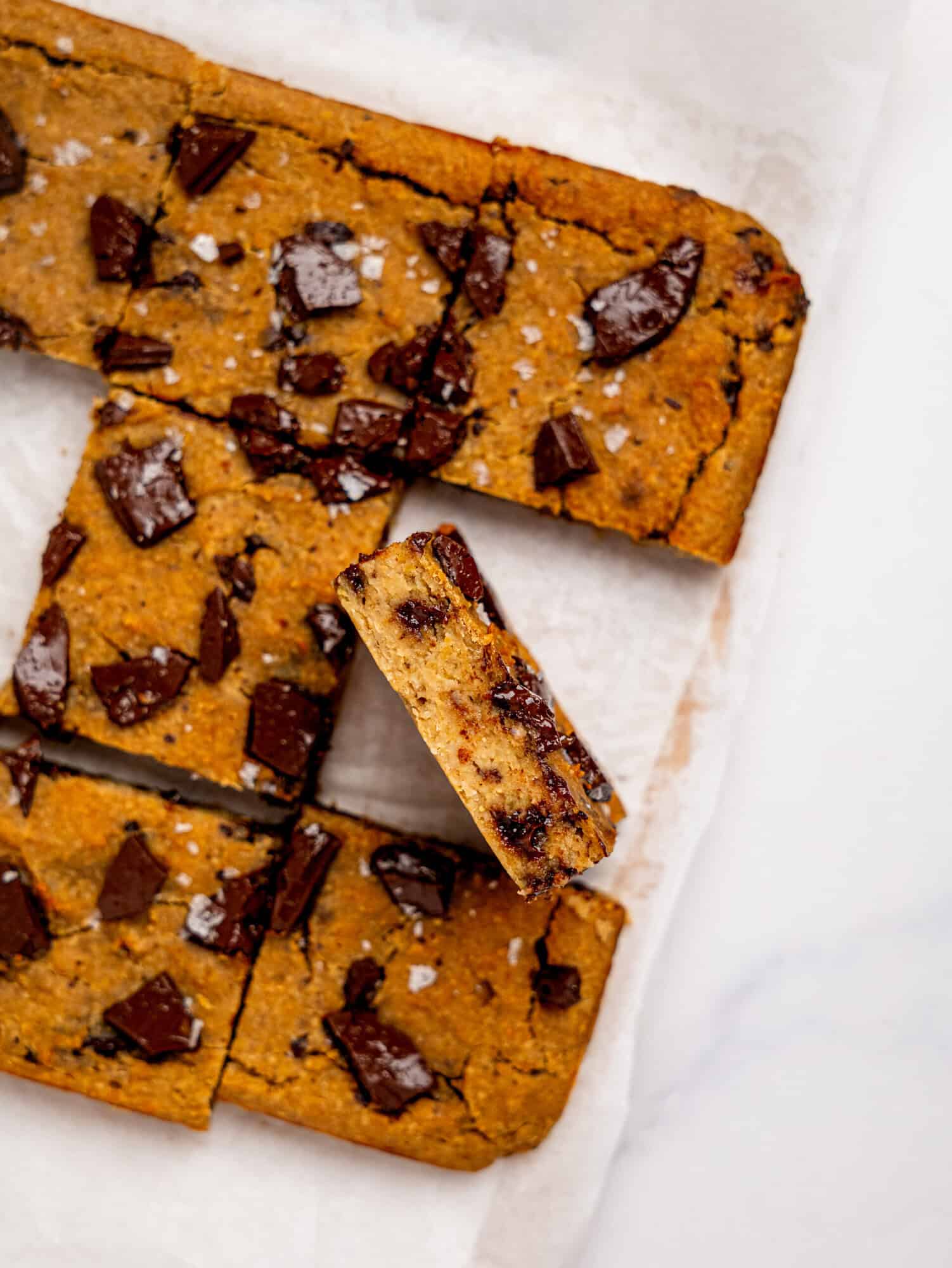 Bird's eye view of chickpea cookie bars on a sheet of white parchment paper, one is turned on its side to reveal the fudgy texture.