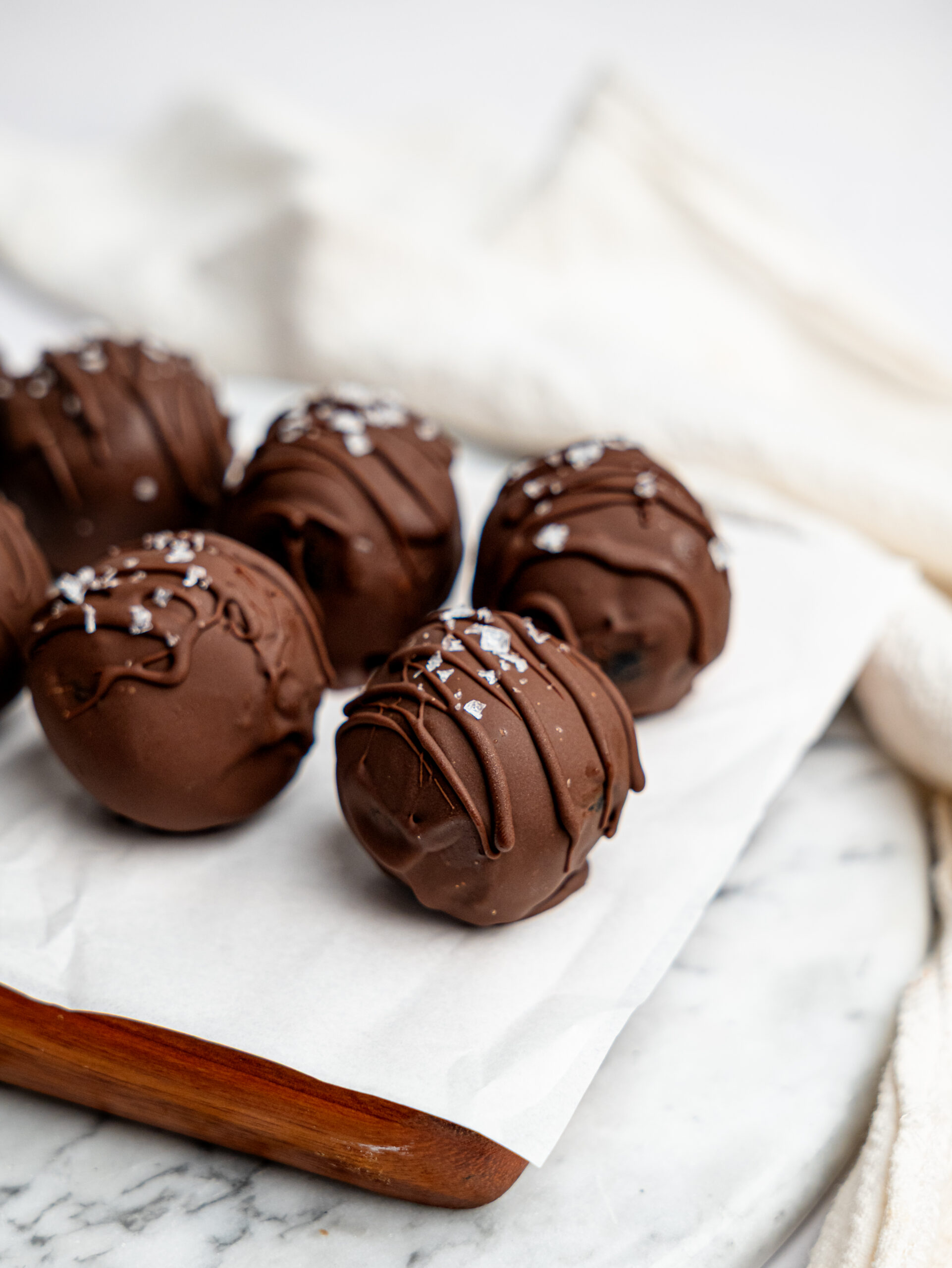 Single chocolate-coated brownie bite topped with flaky sea salt on a wooden tray lined with parchment paper.