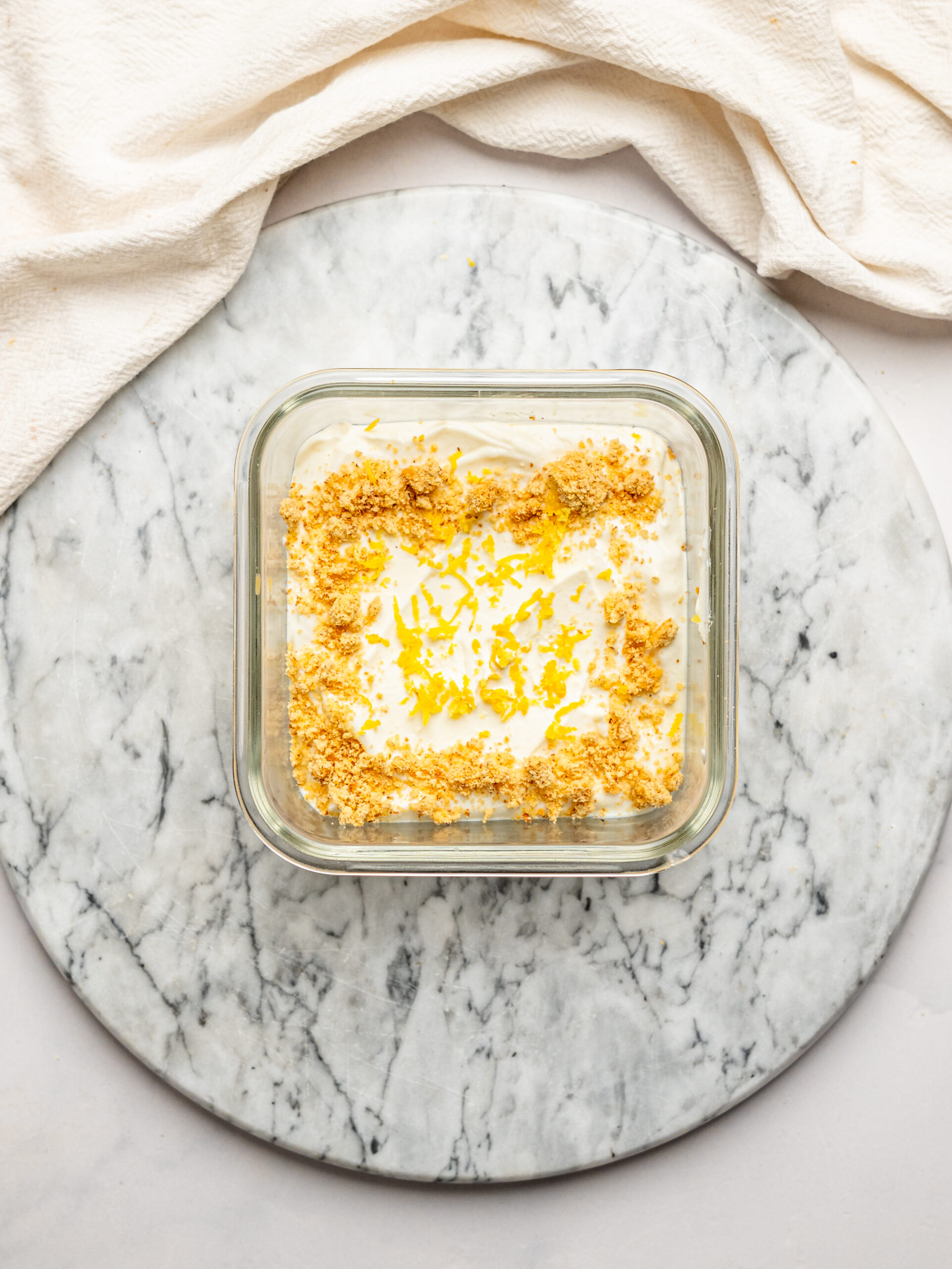 Overhead view of lemon cheesecake overnight Weet-Bix topped with biscuit crumbs and lemon zest, presented on a marble board with a draped cloth background.