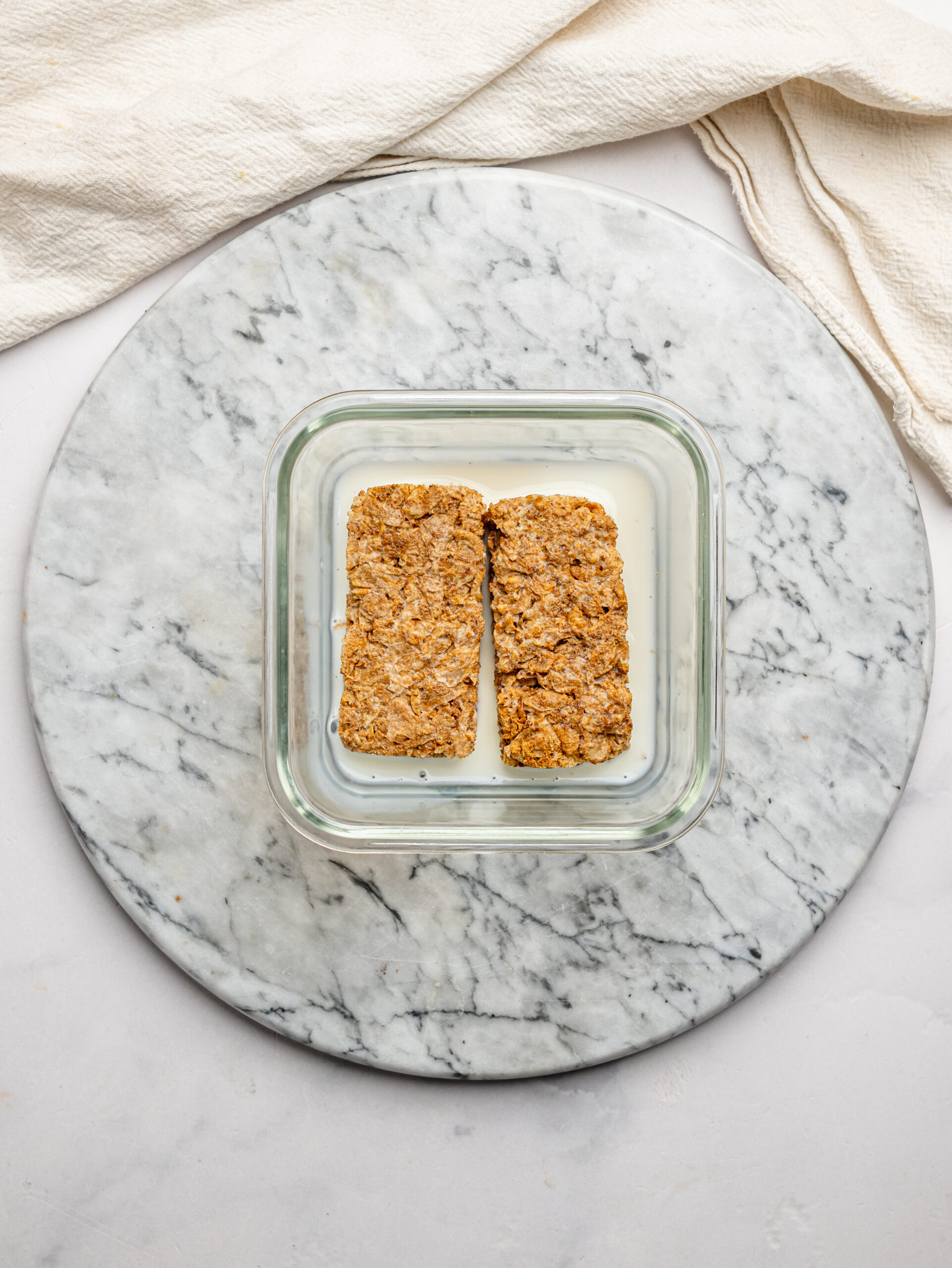 Two Weetbix biscuits soaking in milk in a glass container before being crushed and mixed.