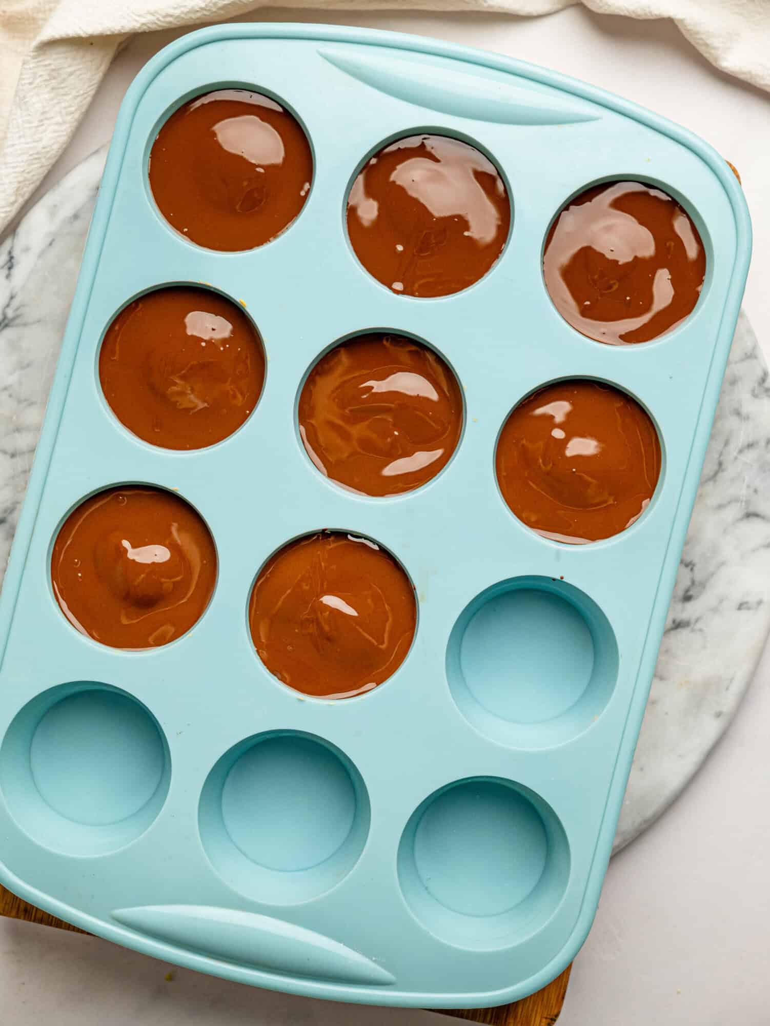 Silicone muffin tray filled with protein peanut butter cups topped with a smooth layer of melted dark chocolate, ready to chill until set.