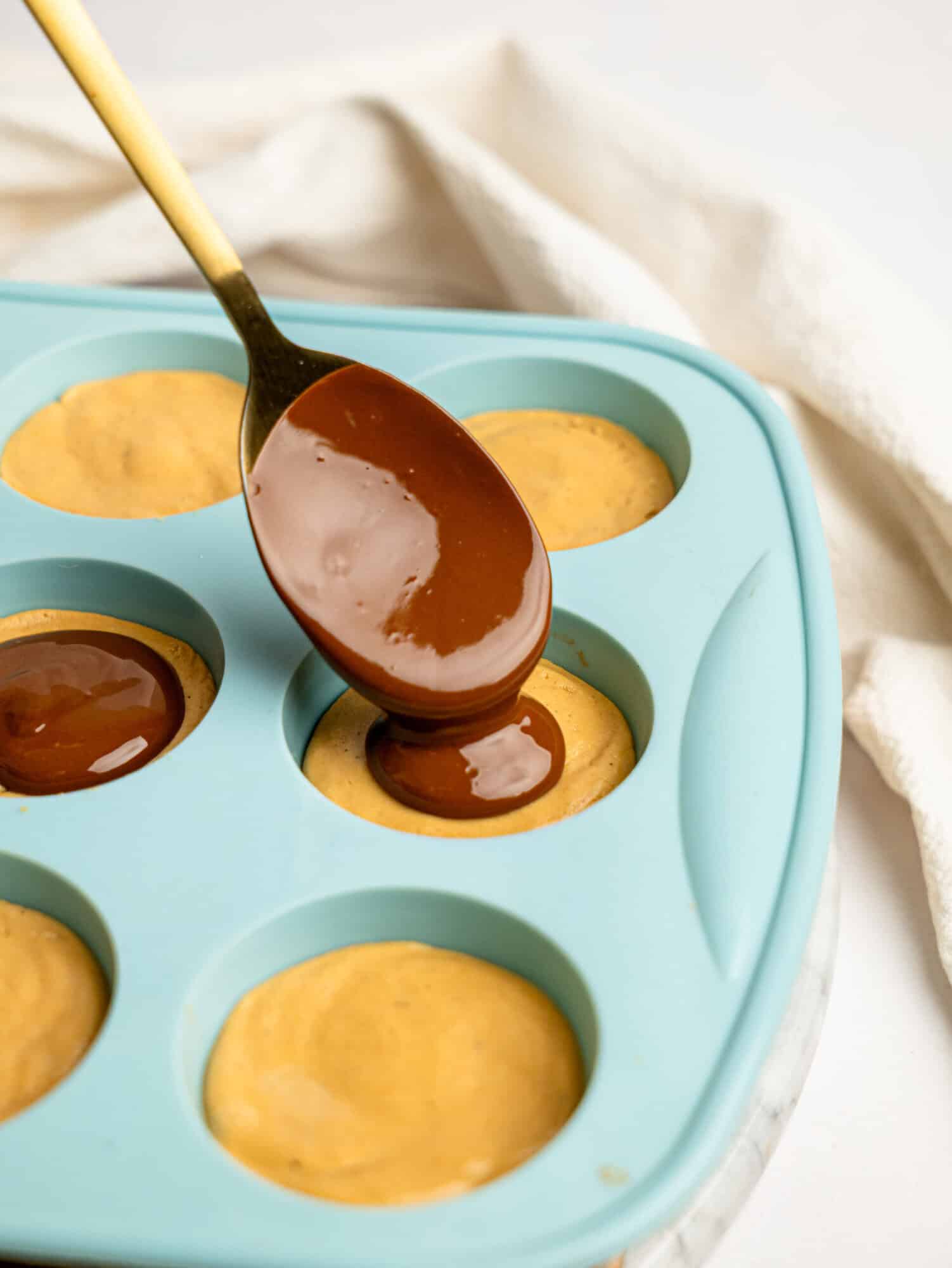 Spoon pouring melted dark chocolate over peanut butter filling in a silicone muffin tray while assembling homemade protein peanut butter cups.