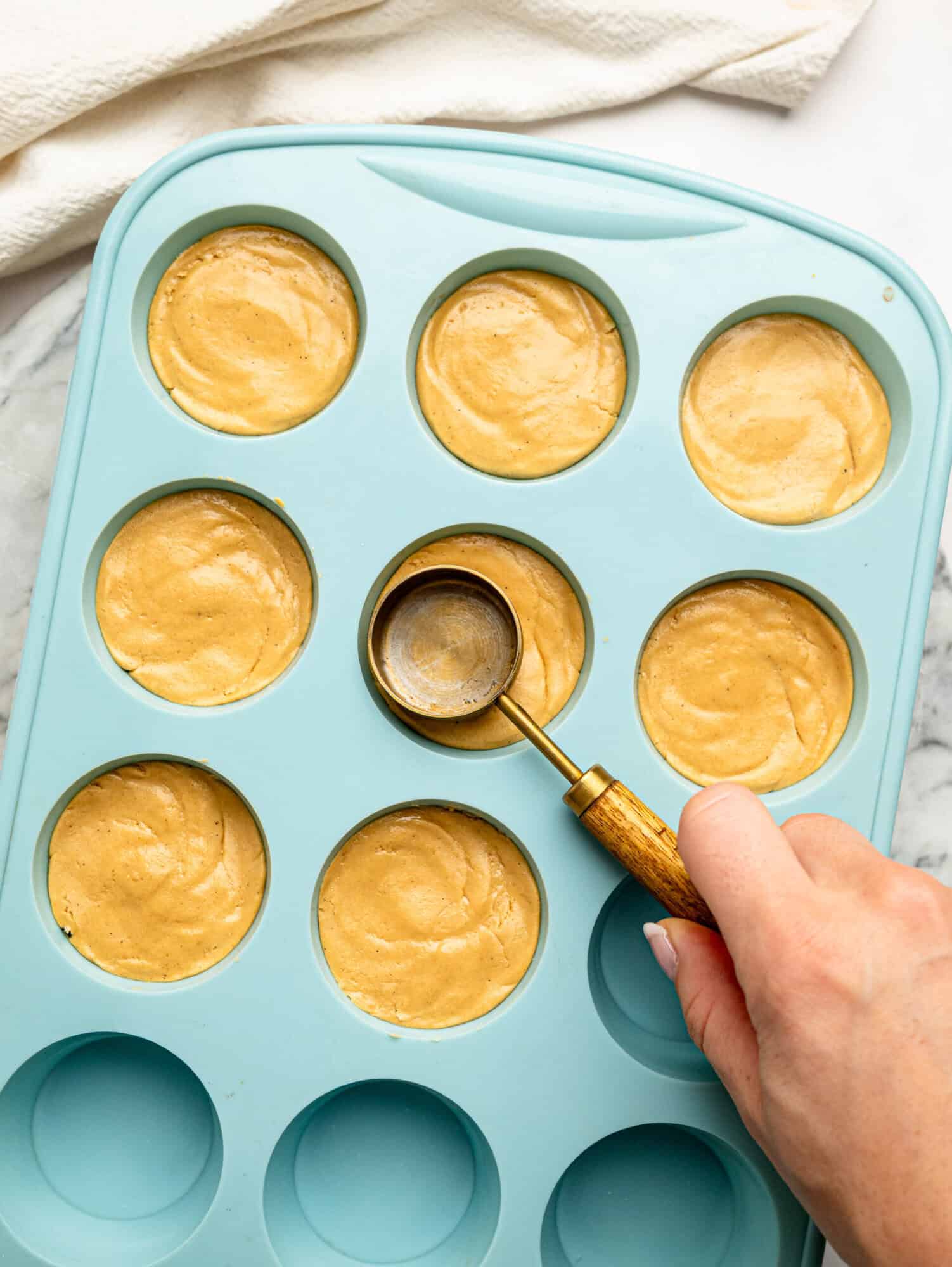 Hand using the back of a measuring spoon to press the peanut butter protein mixture into a silicone muffin tray to form the cup bases.