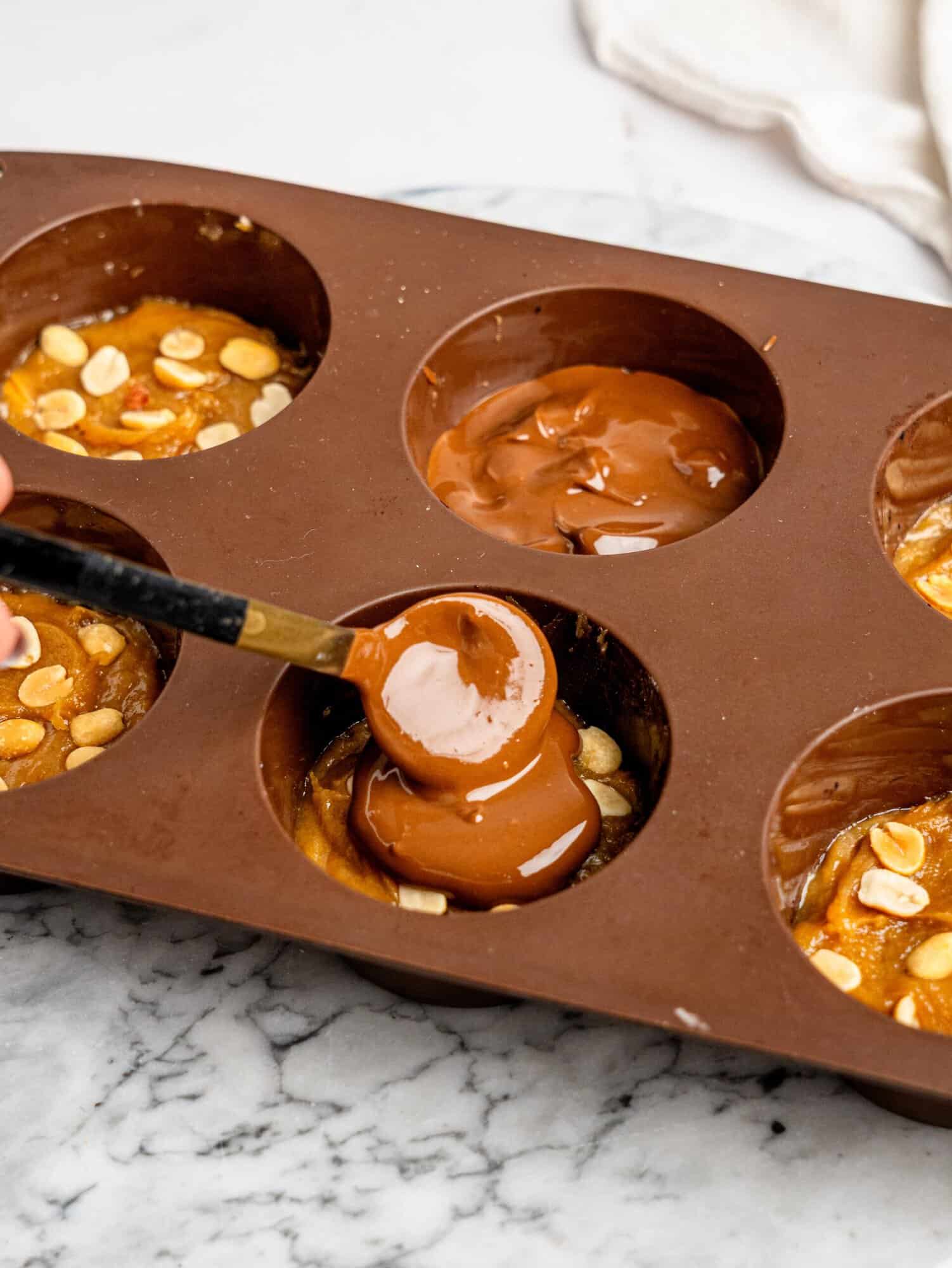 Melted chocolate being spooned over the peanut butter and peanut layer of homemade Snickers Weetbix Cups.