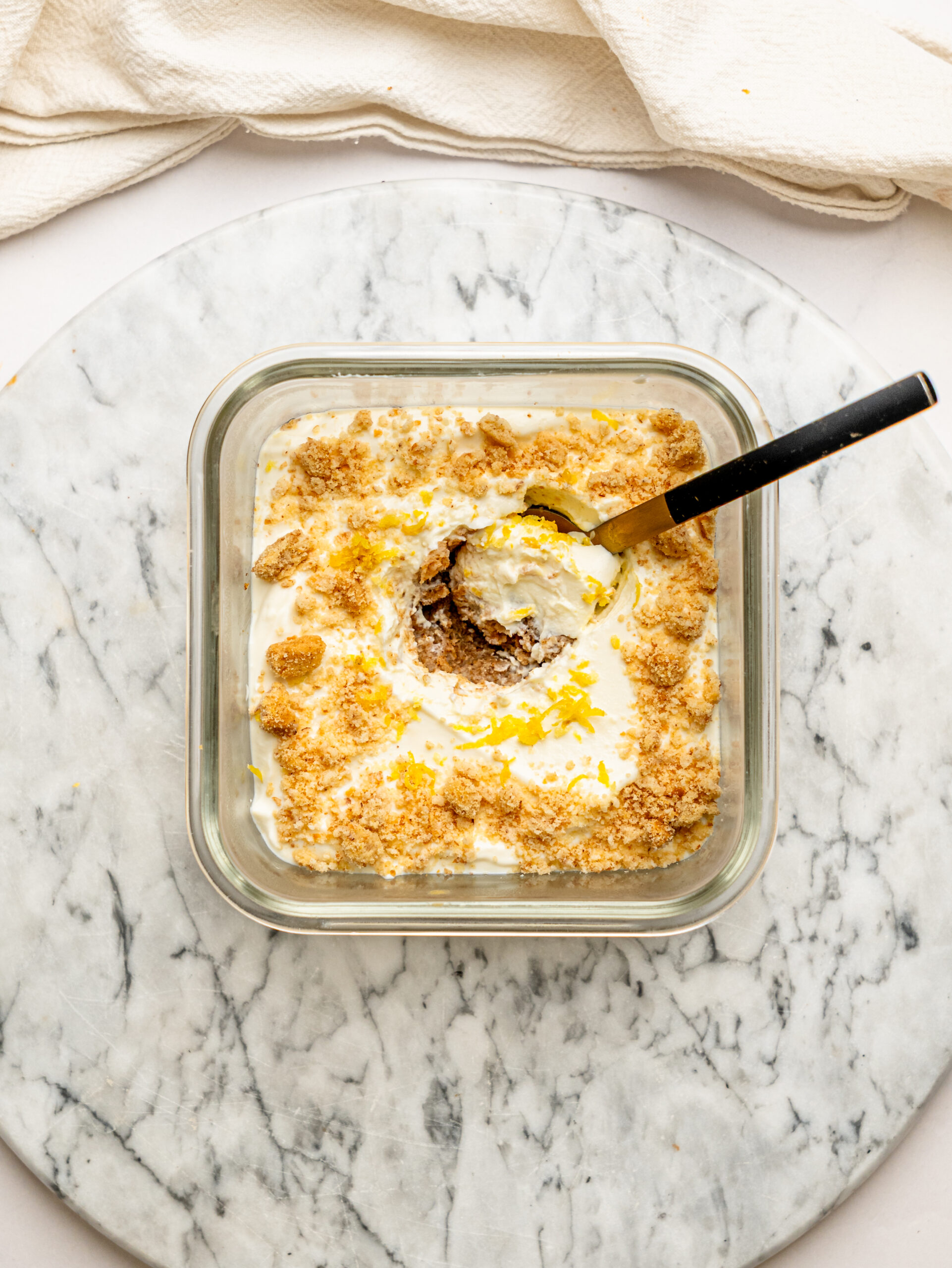 Lemon cheesecake overnight Weet-Bix in a glass meal prep container topped with crushed biscuit and fresh lemon zest, with a spoon scooping out a creamy serving.