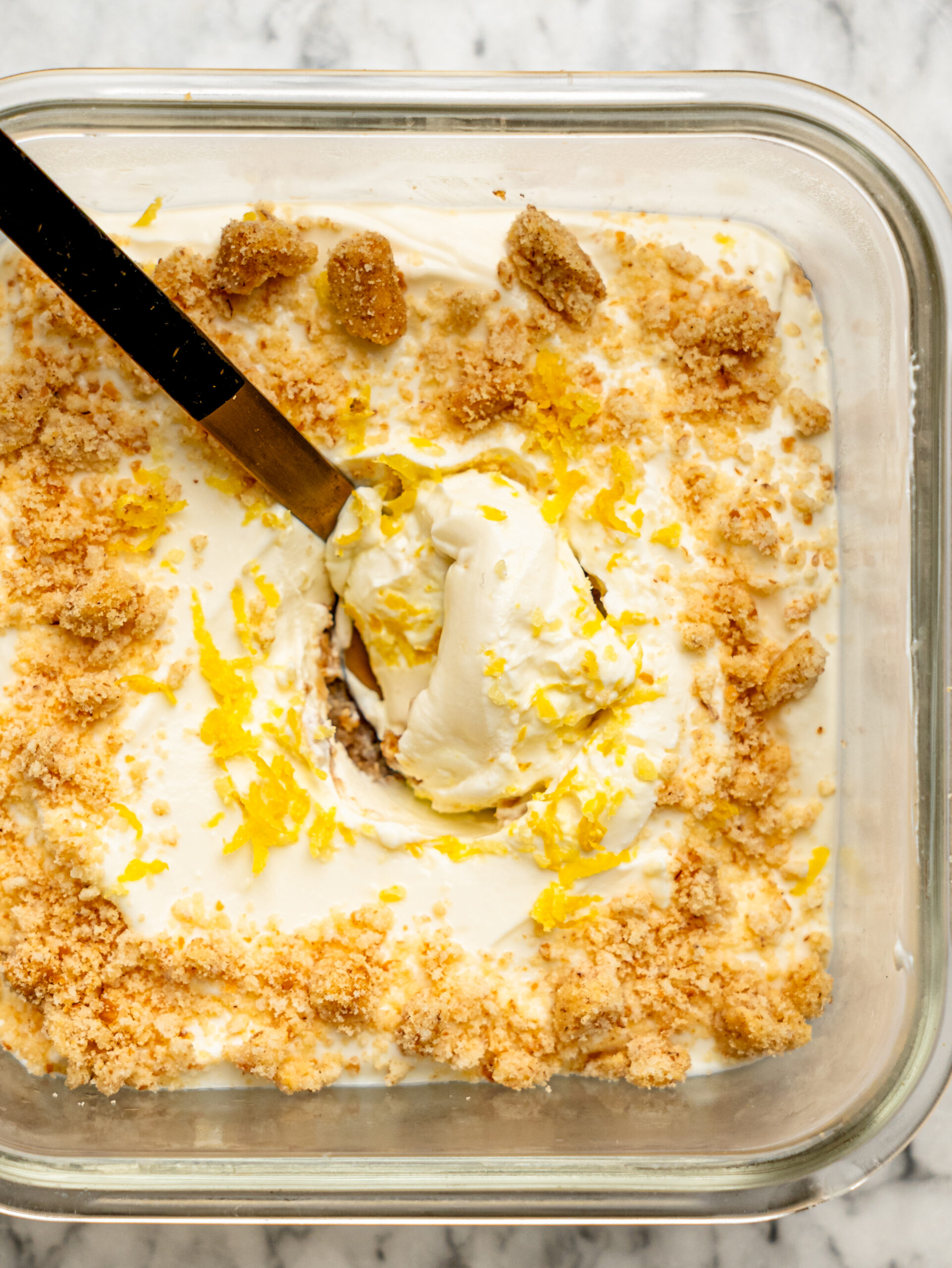 Close-up overhead view of lemon cheesecake overnight Weet-Bix with a spoon lifting the creamy lemon cheesecake layer, finished with biscuit crumbs and lemon zest.