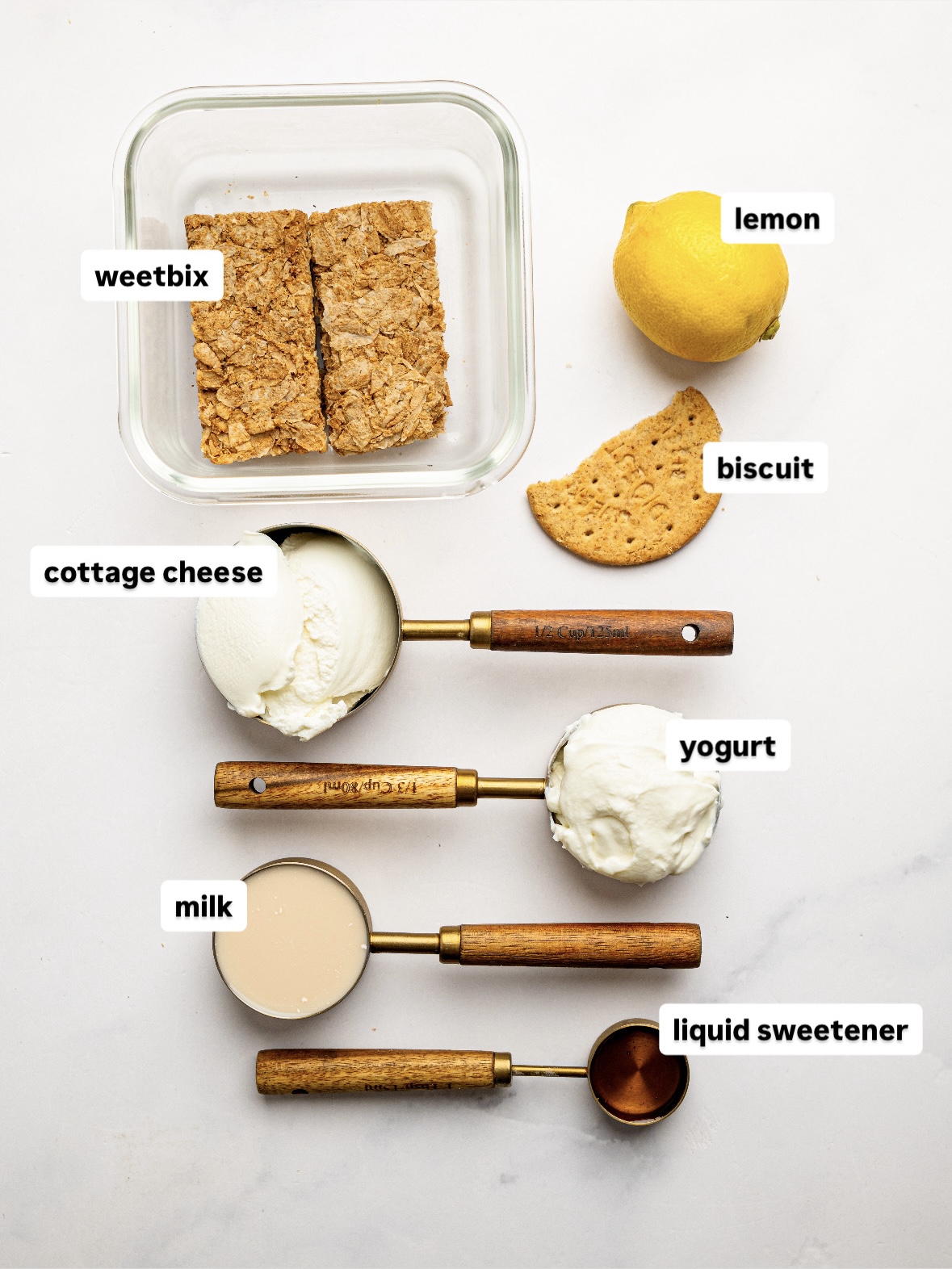 Ingredients for lemon cheesecake overnight Weet-Bix arranged on a white surface, including Weet-Bix biscuits, cottage cheese, Greek yogurt, milk, liquid sweetener, a lemon, and a biscuit for topping.