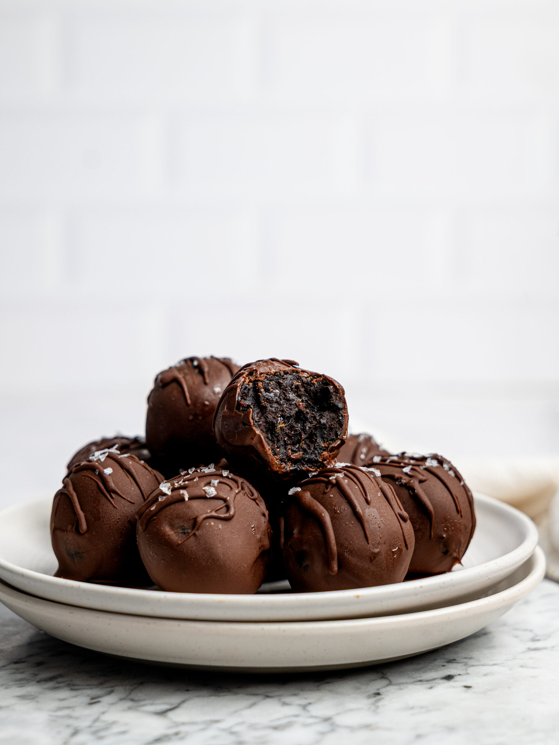 Stack of chocolate-covered no-bake brownie bites on a white plate, topped with flaky sea salt and one brownie bite bitten open.