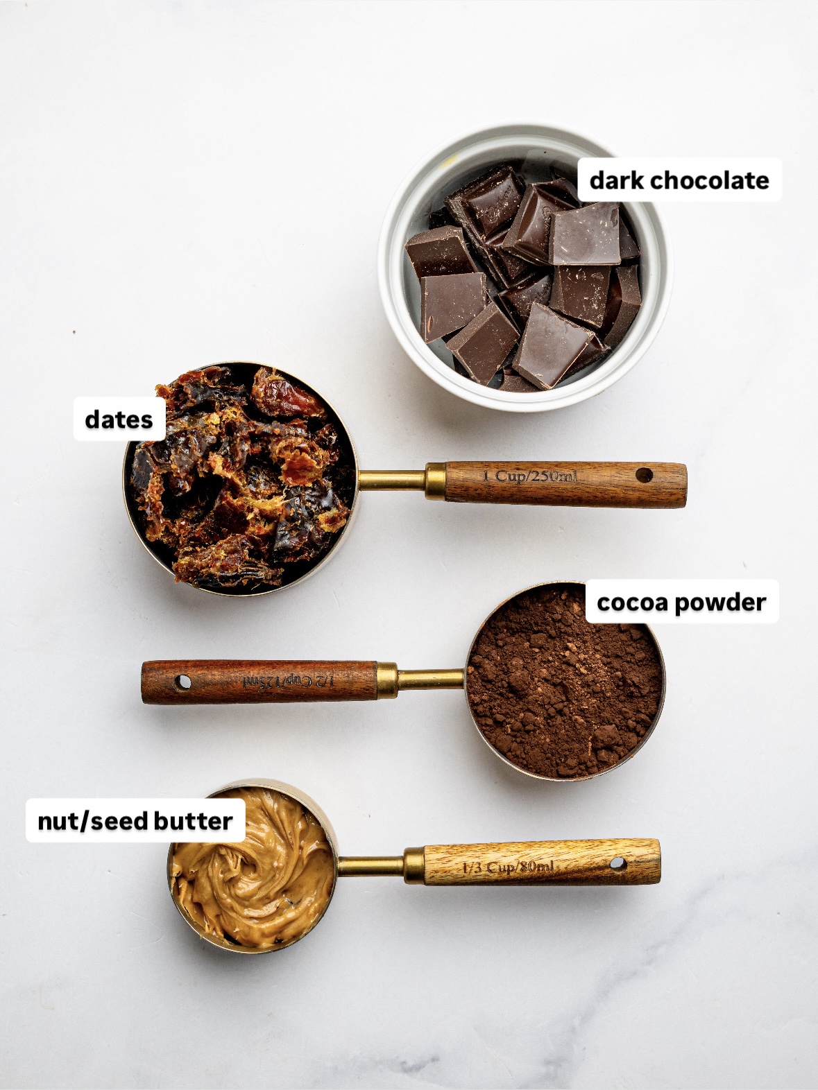 Ingredients for No-Bake Brownie Bites measured out in bowls and measuring cups, including dates, cocoa powder, nut butter, and dark chocolate.
