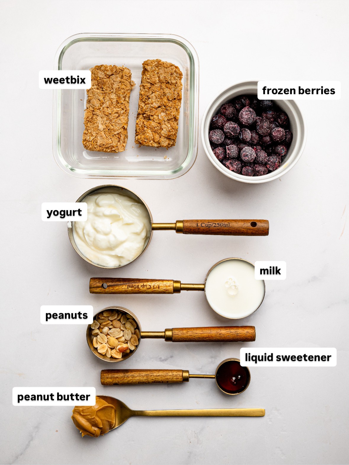 Ingredients for PB&J Overnight Weetbix including Weetbix, Greek yogurt, milk, peanut butter, honey, frozen berries, and peanuts arranged on a white surface.