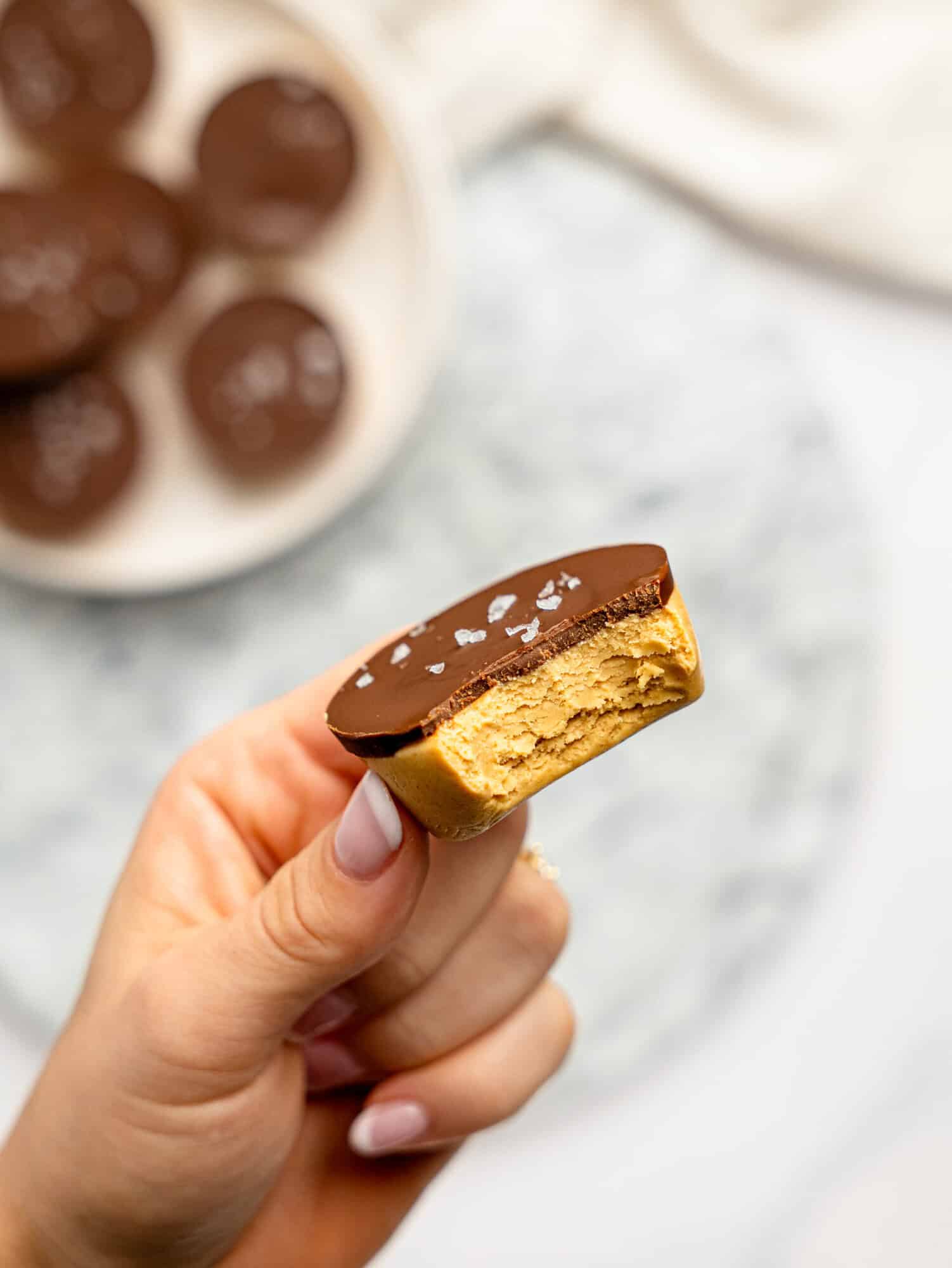 Hand holding a bitten protein peanut butter cup, revealing the thick, fudgy peanut butter filling beneath a smooth dark chocolate topping with a sprinkle of flaky sea salt.