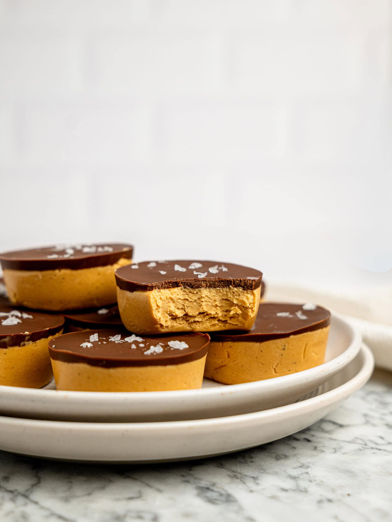 Homemade protein peanut butter cups topped with flaky sea salt on stacked white plates, featuring one cup with a bite taken out to reveal the soft peanut butter filling.