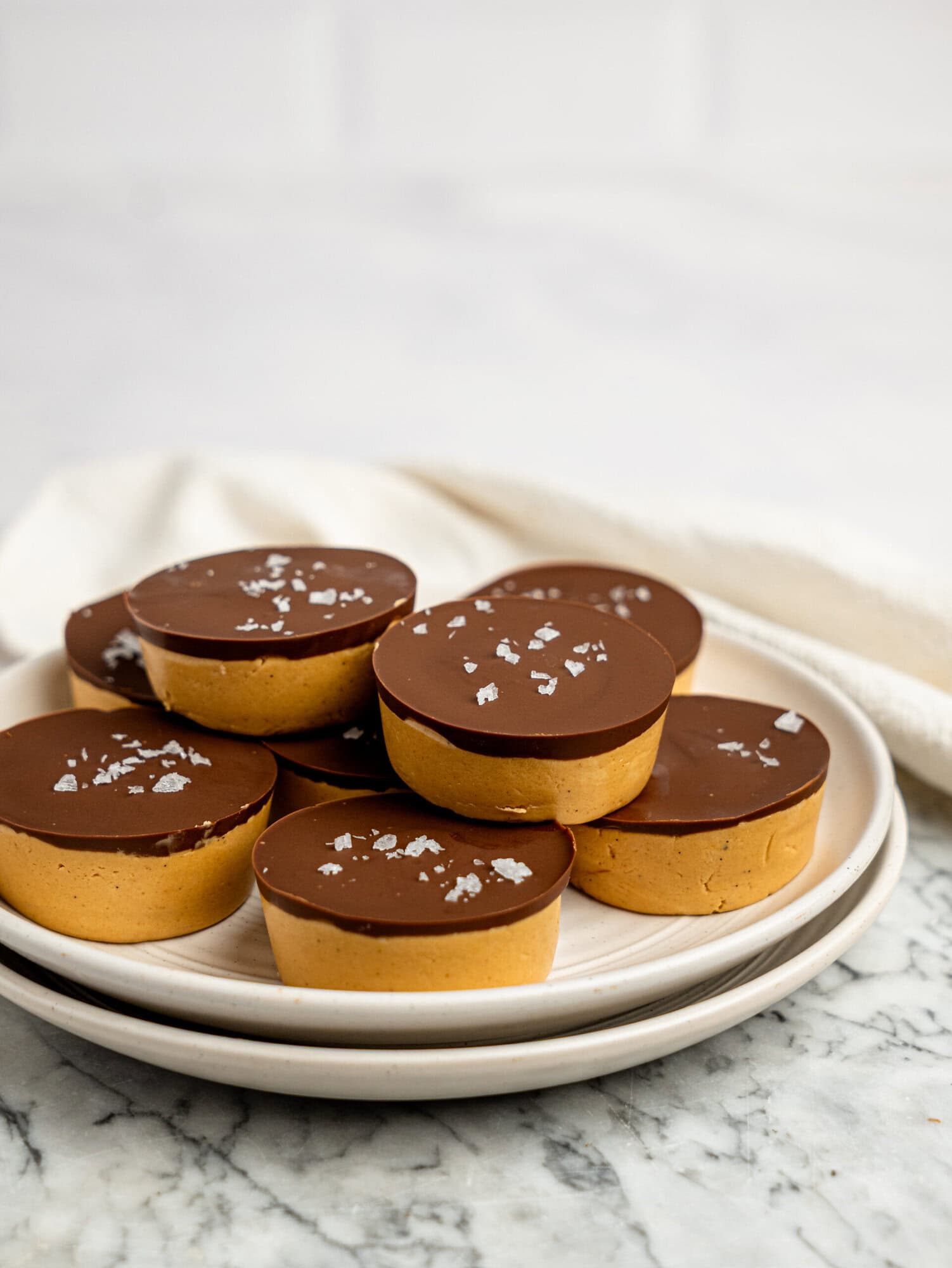 Plate of no-bake protein peanut butter cups topped with flaky sea salt, finished with a glossy dark chocolate coating and creamy peanut butter filling.