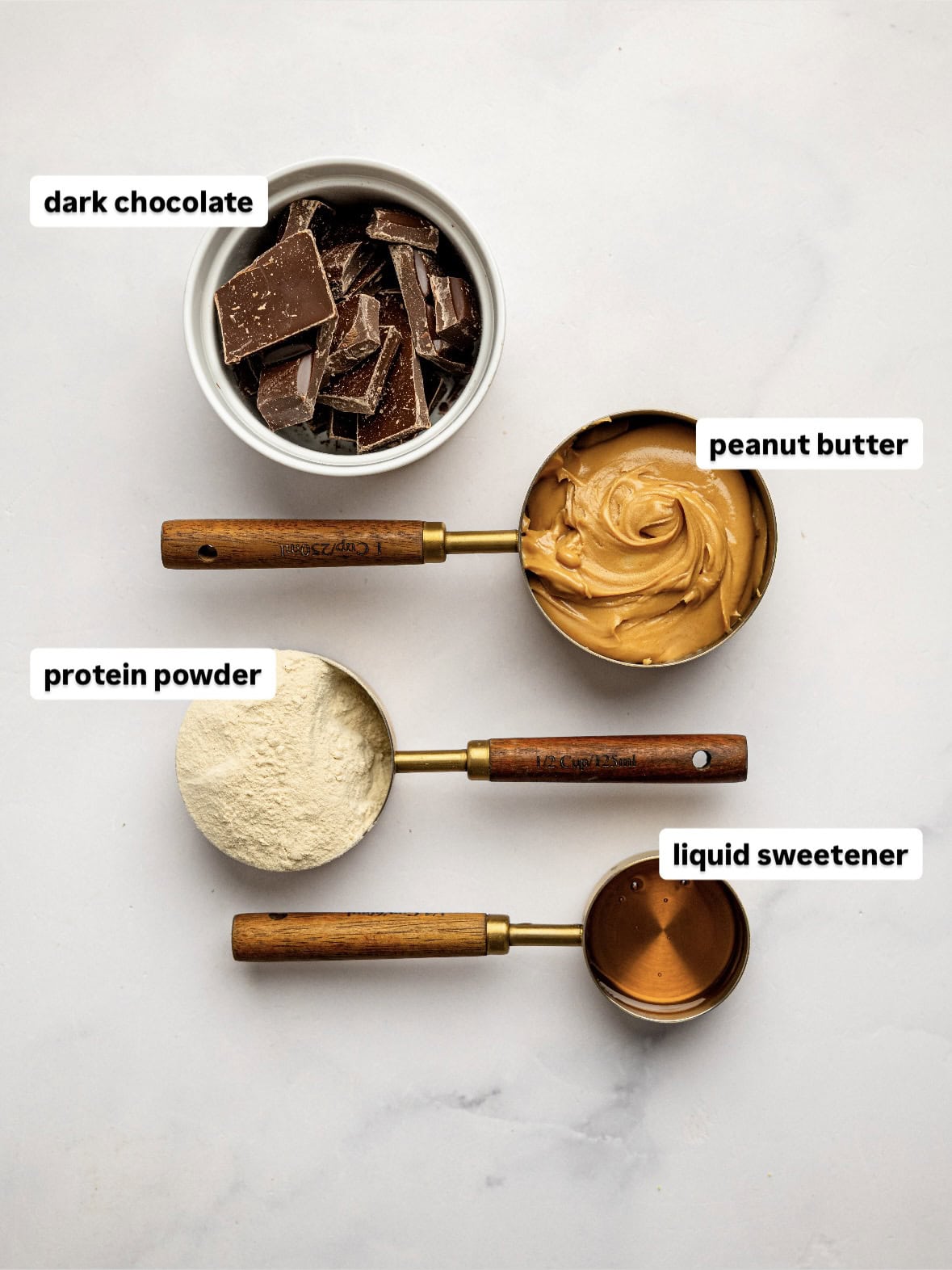 Ingredients for protein peanut butter cups arranged on a white surface, including chopped dark chocolate, smooth peanut butter, vanilla protein powder, and liquid sweetener in measuring cups and bowls.