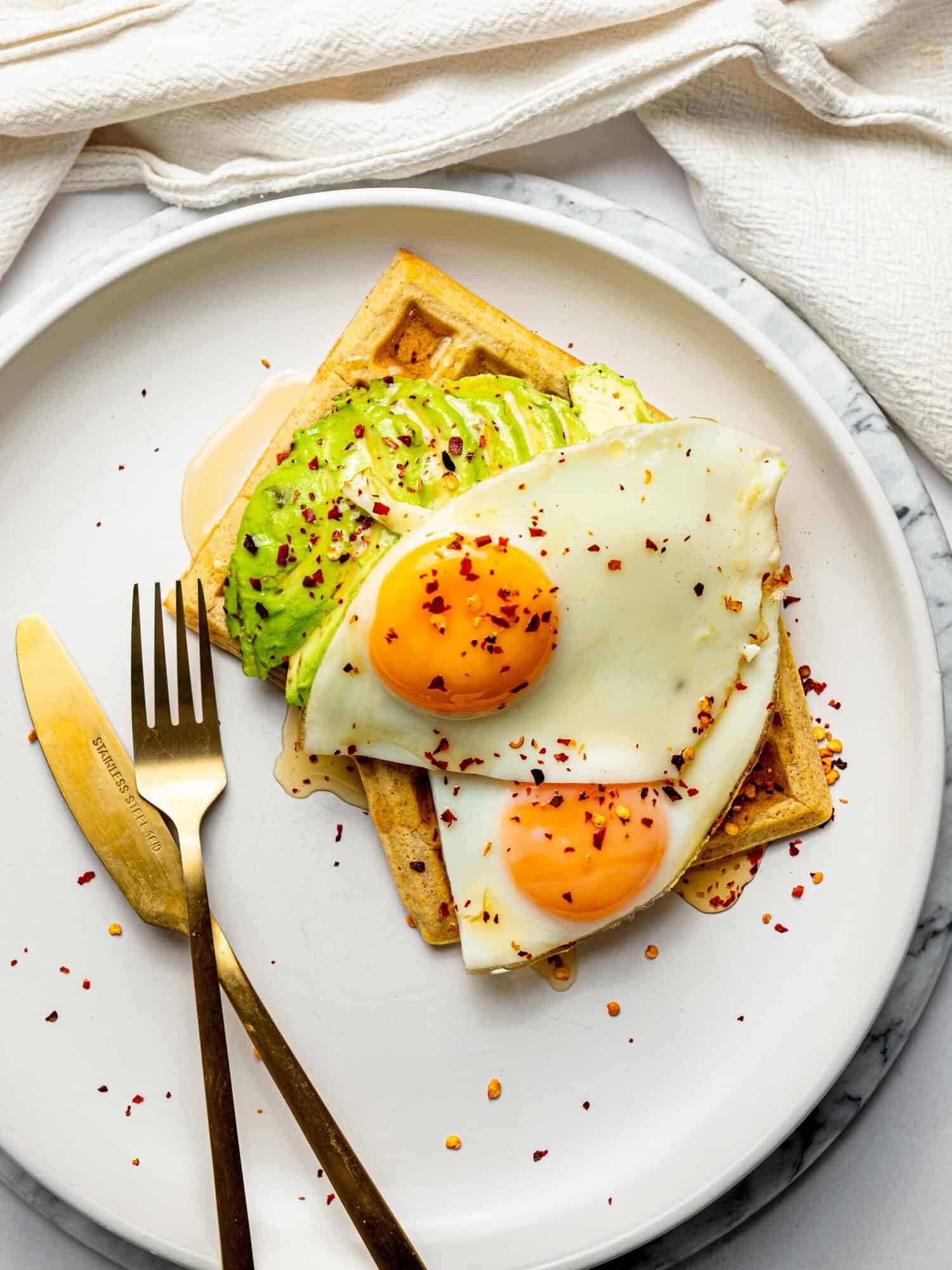 Gluten-free waffles topped with avocado, fried eggs, and chili flakes for a savory breakfast.