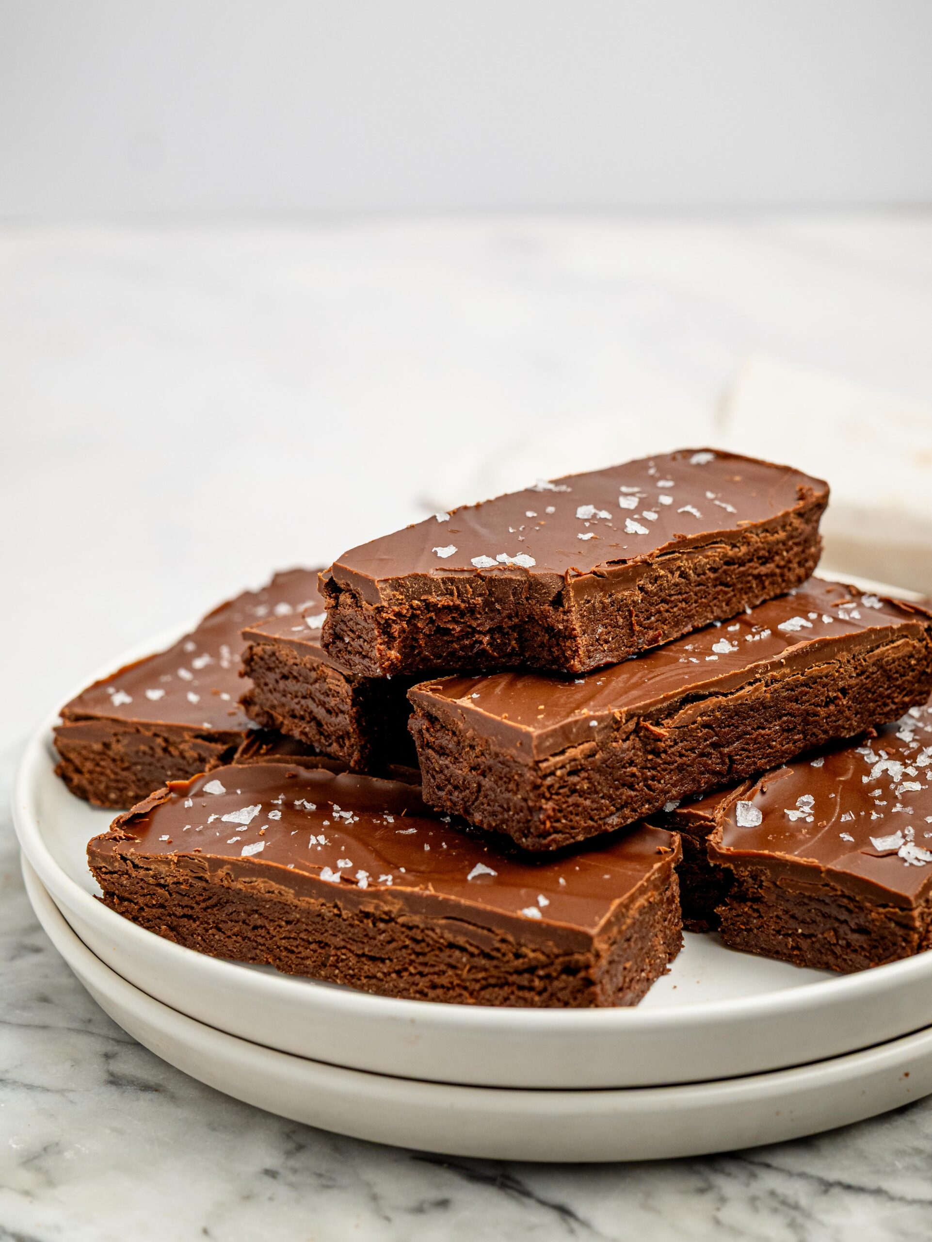 Chocolate fudge protein bars topped with a smooth chocolate layer and flaky sea salt stacked on a white plate.