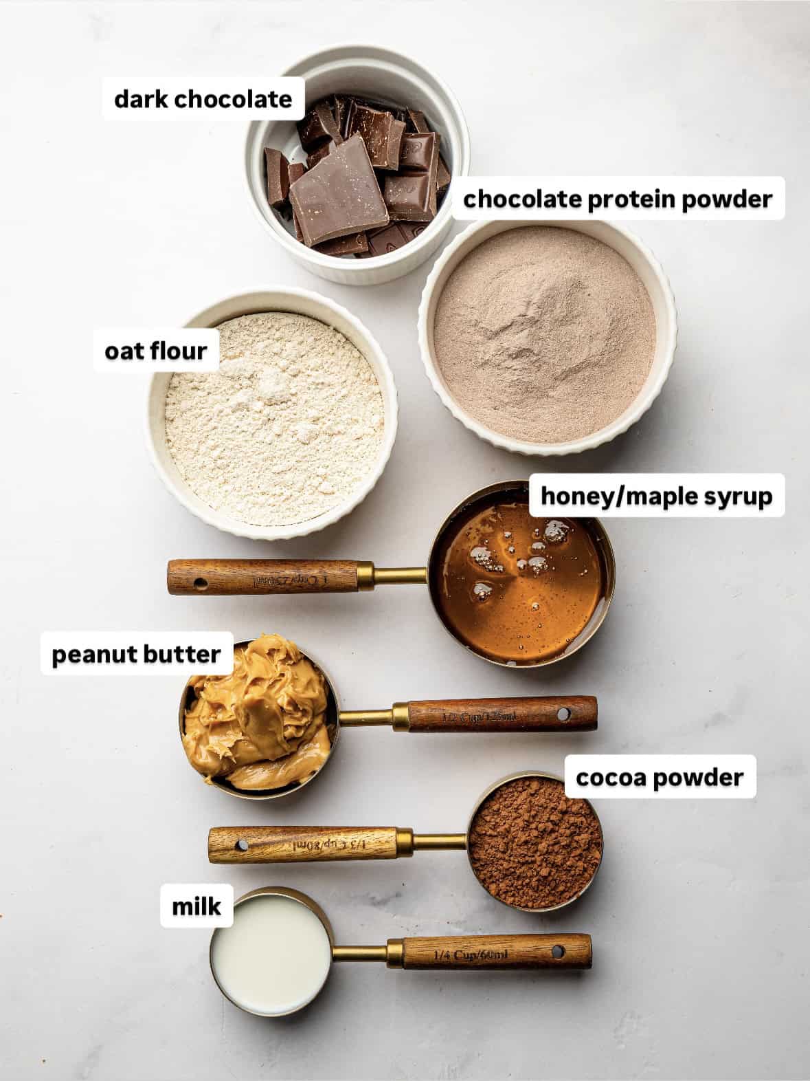Ingredients for homemade chocolate protein bars including chocolate protein powder, oat flour, peanut butter, honey, cocoa powder, milk, and dark chocolate.